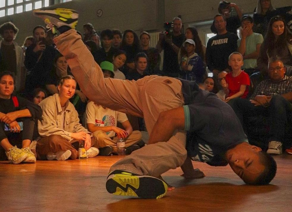 Un ballerino di breakdance con pantaloni beige e camicia scura esegue un freeze su un palco davanti a un pubblico seduto.
