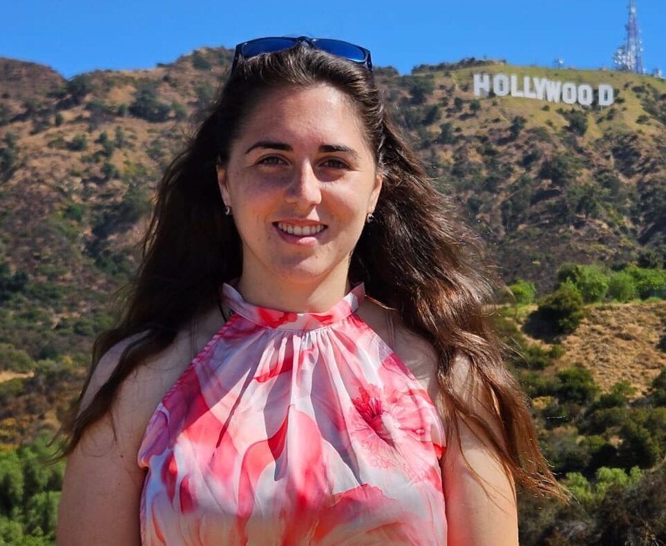 Una donna con una maglietta rosa a fantasia sorride davanti alla scritta Hollywood su una collina soleggiata.