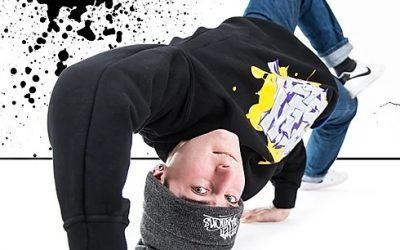 Un uomo esegue una presa a una mano in stile breakdance su un pavimento bianco da studio, indossando una felpa nera e jeans.