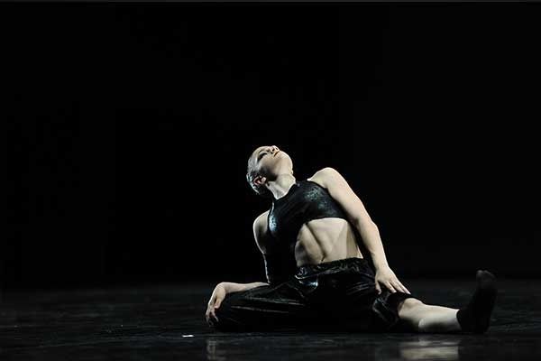 Ballerina in abito nero seduta sul pavimento contro uno sfondo scuro, reclinata all'indietro in una posa drammatica.