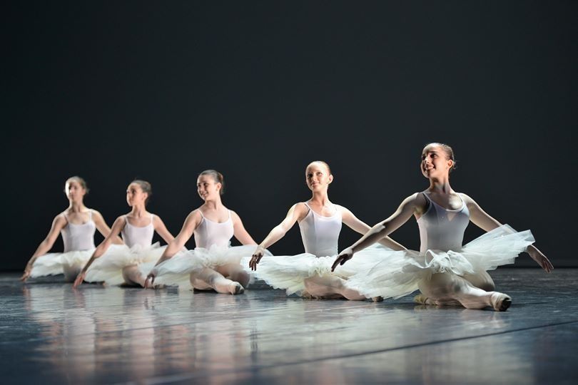 Cinque ballerine in tutù bianchi sedute in fila su un palcoscenico buio, tenendosi per mano