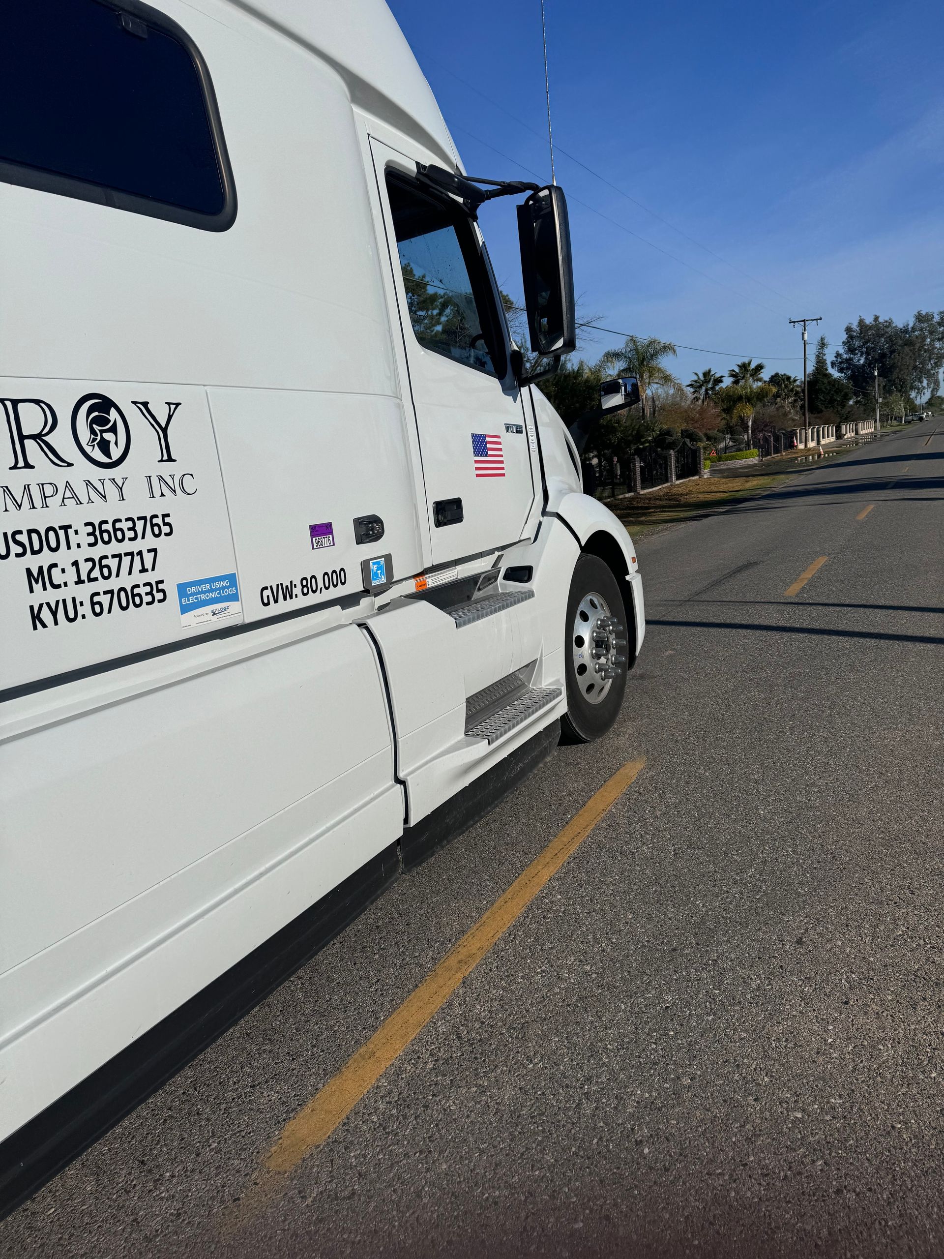 White semi-truck parked on asphalt road, side view. Truck has company name and U.S. flag on the side.