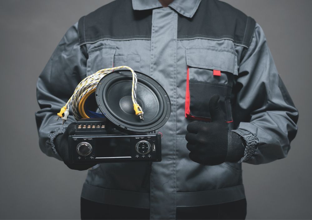 Fitter in grey overalls holding a car stereo, a speaker and wiring, with a thumbs up