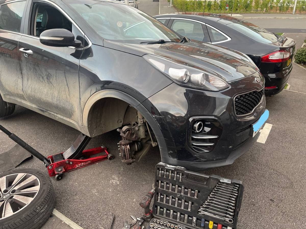Black Kia Sportage with its front wheel removed and a socket set beside it in a parking bay