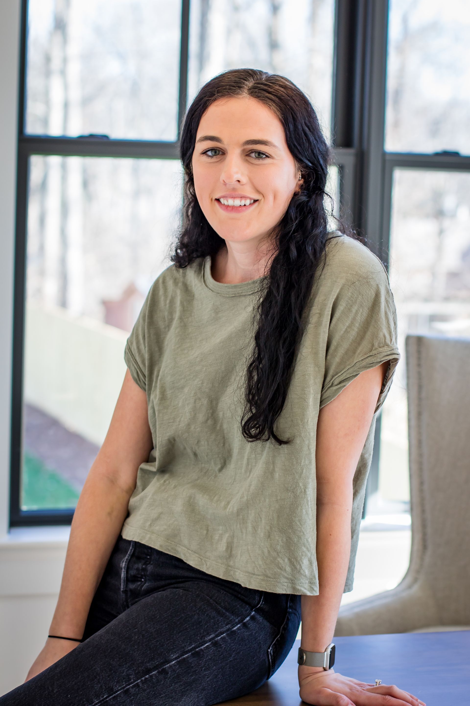 Woman with dark hair, smiling, wearing a green shirt and dark jeans, leaning on a table near a window.