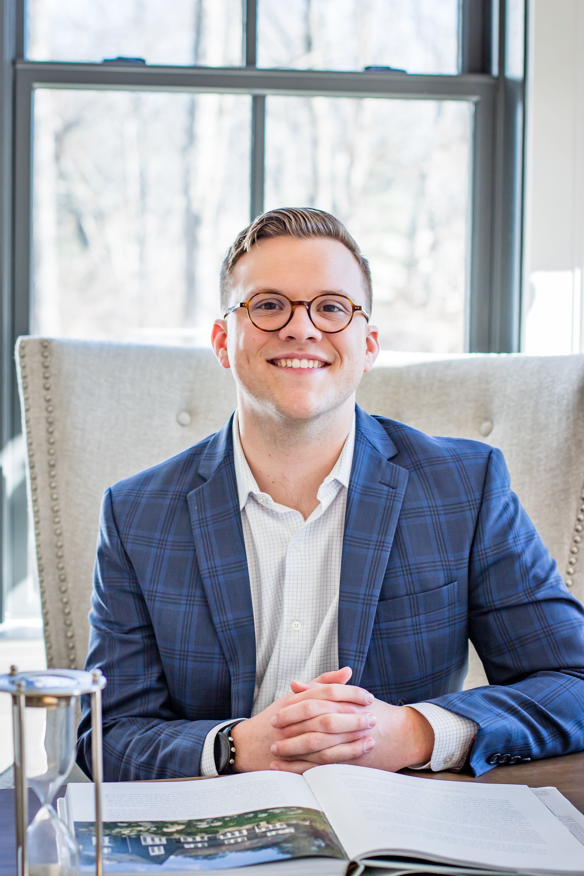Man in blue blazer smiles, seated at a desk near a window, with an open book and hourglass.
