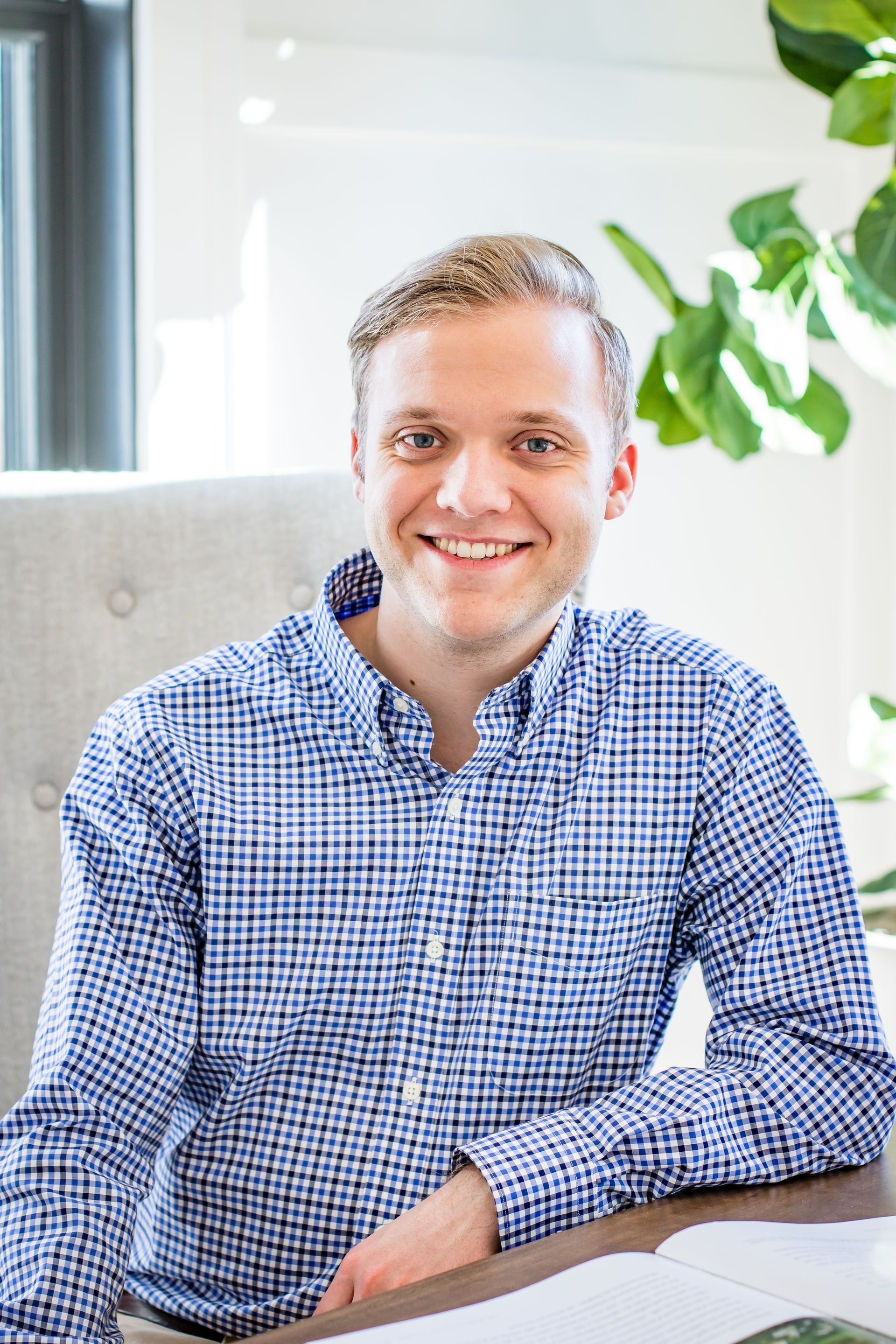 Man in blue checkered shirt smiles, sitting at a desk with greenery in the background.