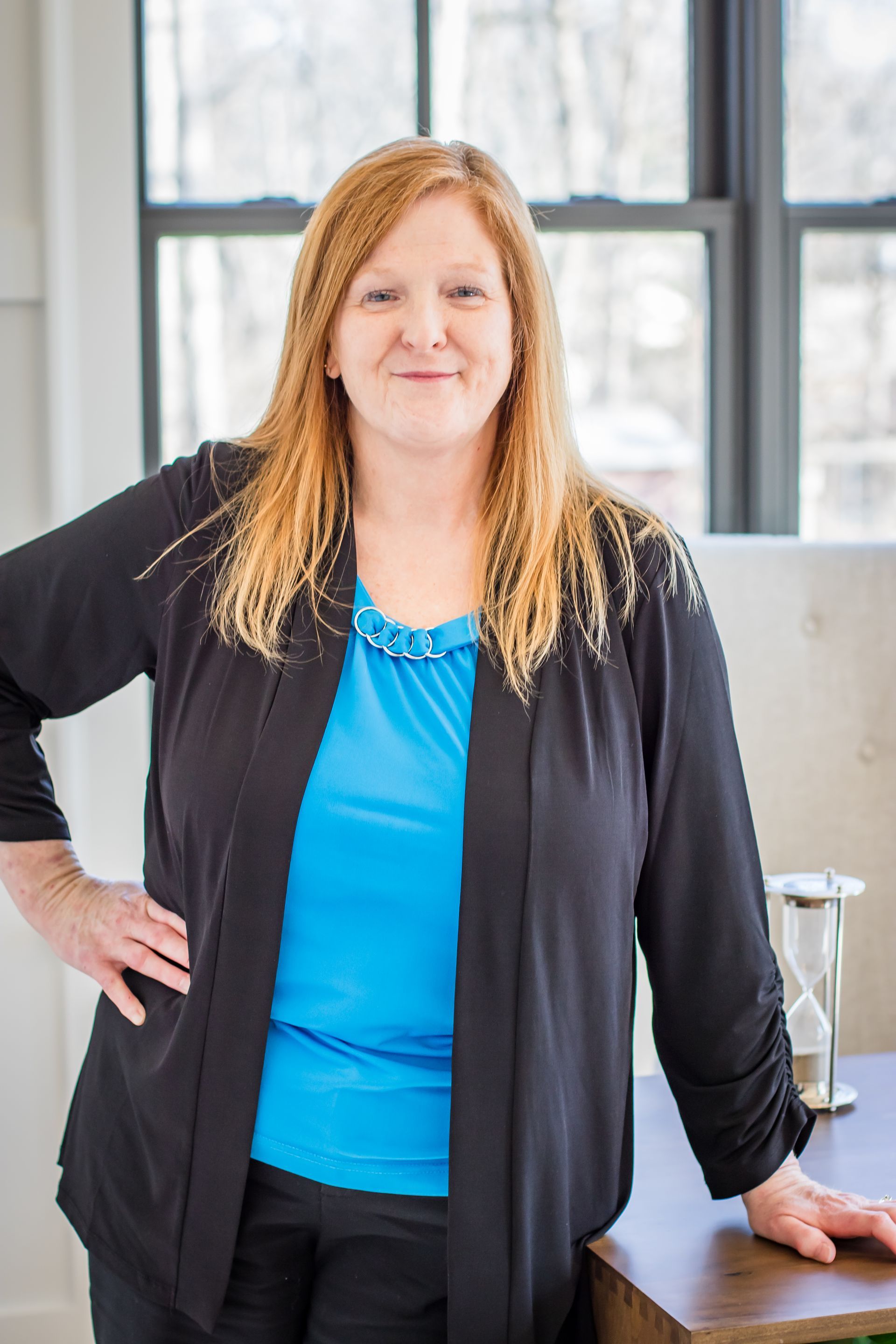 Woman with red hair, blue top, and black jacket, leans on a desk by a window; smiles.