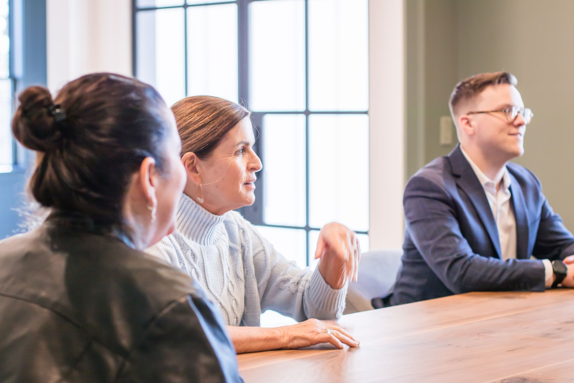 Three people at a table; woman gestures while speaking, man smiles, another woman listens.