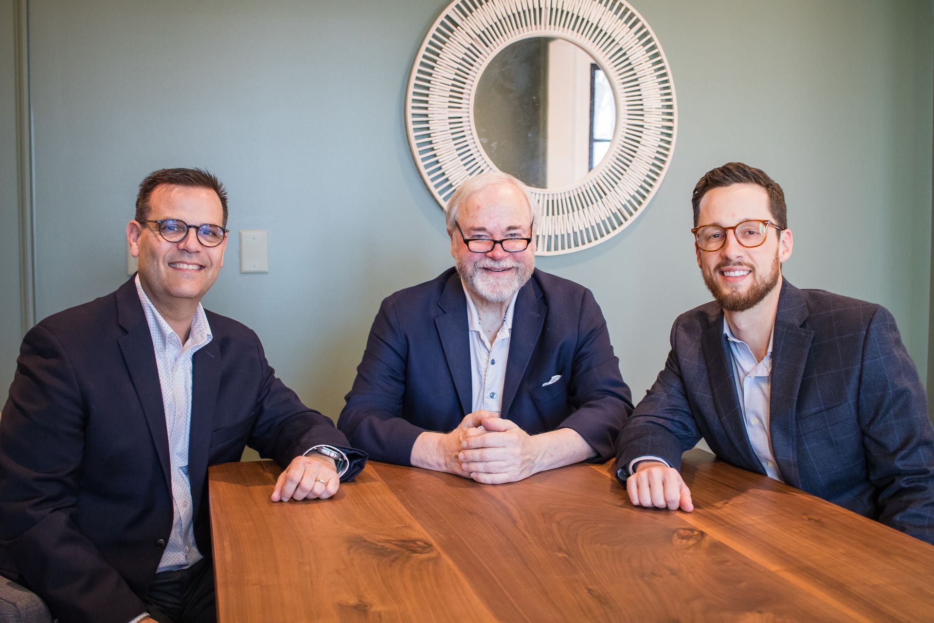 Three men at a wooden table, smiling; one with glasses sits in the middle, framed mirror on wall.