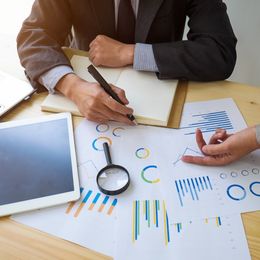 Hands reviewing colorful charts and graphs with a magnifying glass on a desk with a tablet and notebook