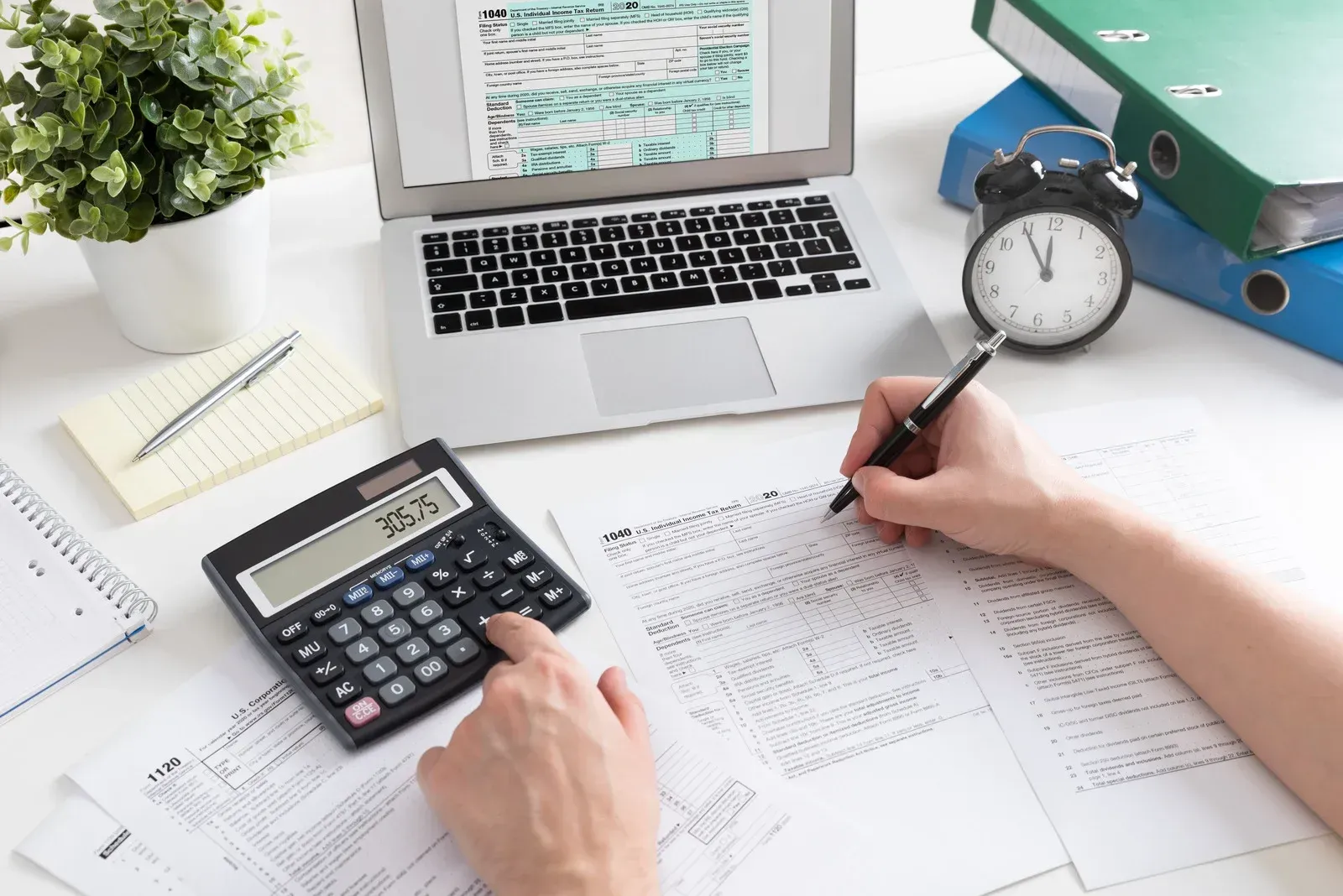 Hands calculating on papers at a desk with laptop, calculator, clock, and office supplies