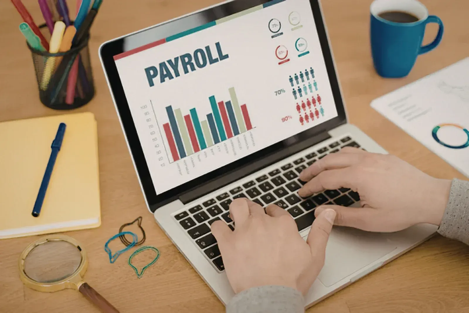 Hands typing on laptop showing payroll charts on a desk with stationery and coffee mug