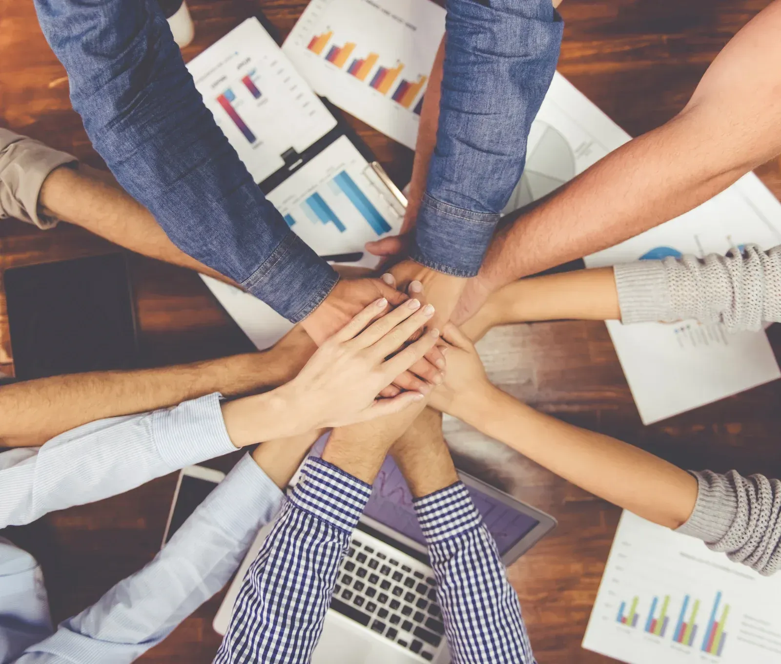 Hands stacked together in a teamwork meeting over charts and a laptop on a table
