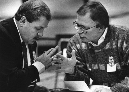 Two men are sitting at a table talking to each other in a black and white photo.