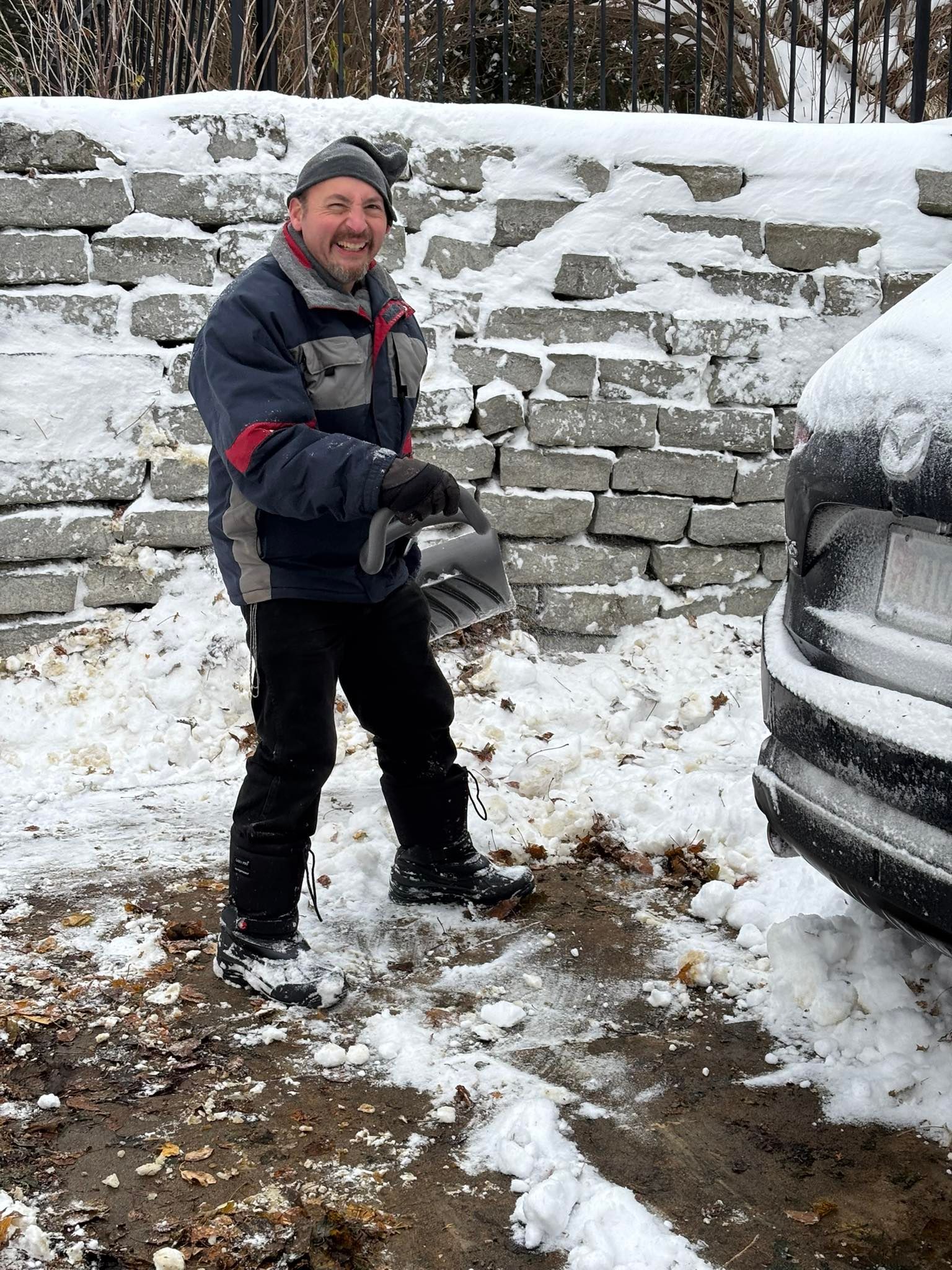 Person in winter clothes standing in snow beside a car and stone wall, smiling and holding a snowball