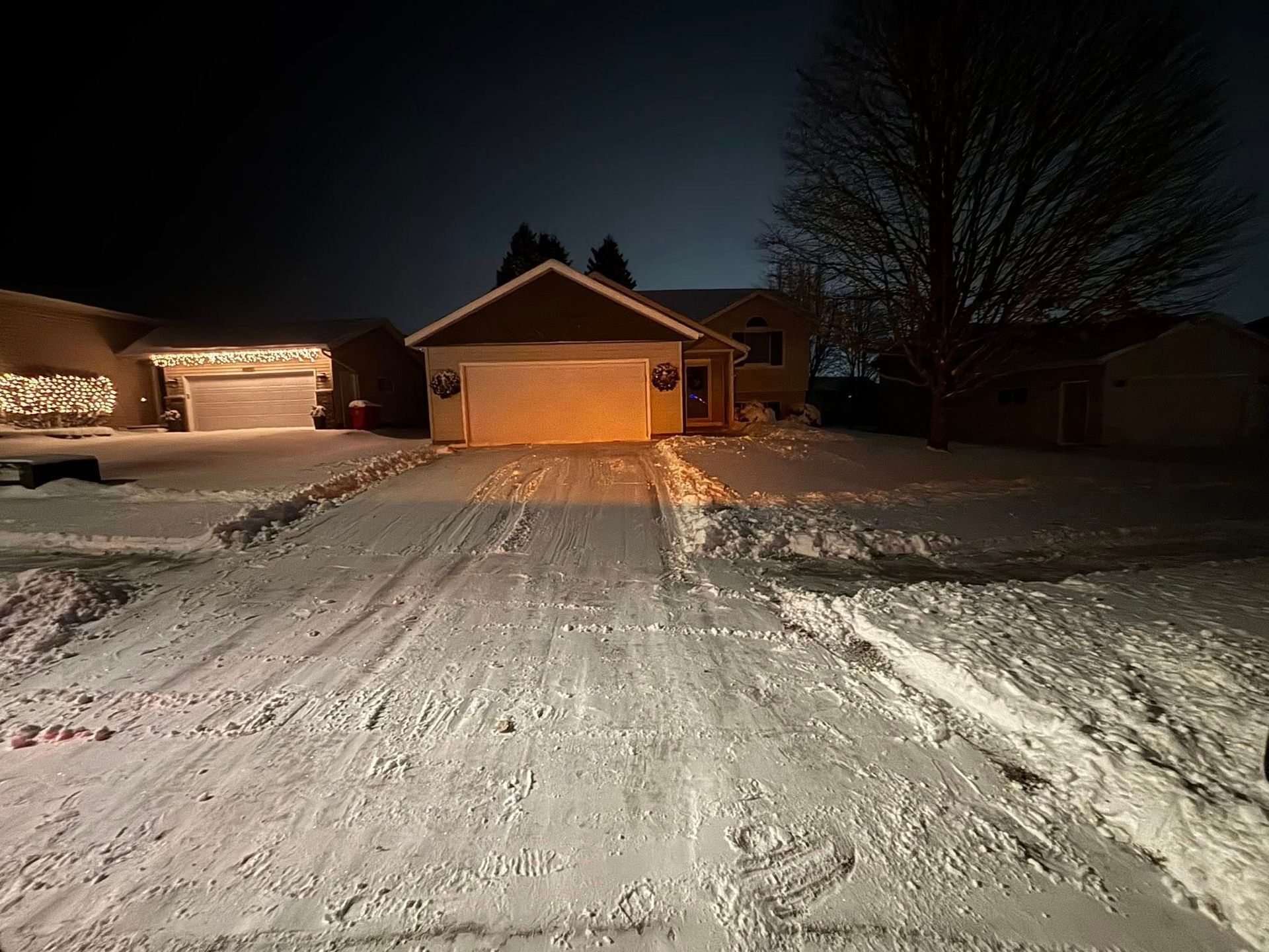 Snow-covered suburban driveway at night, with a lit garage and dark houses under a clear sky