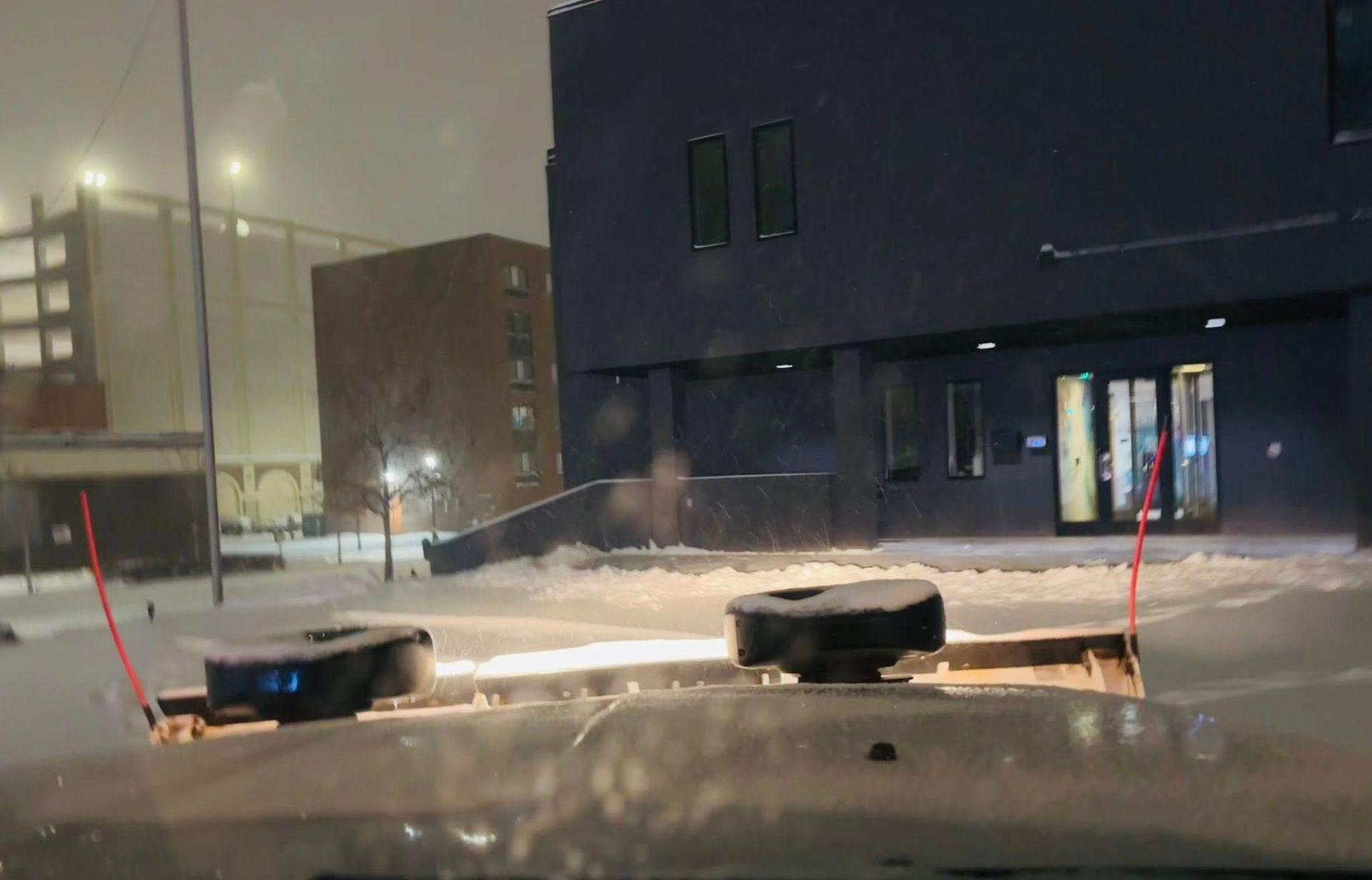 Snowy urban street at night with two black bins near a dark building and parked cars in the background