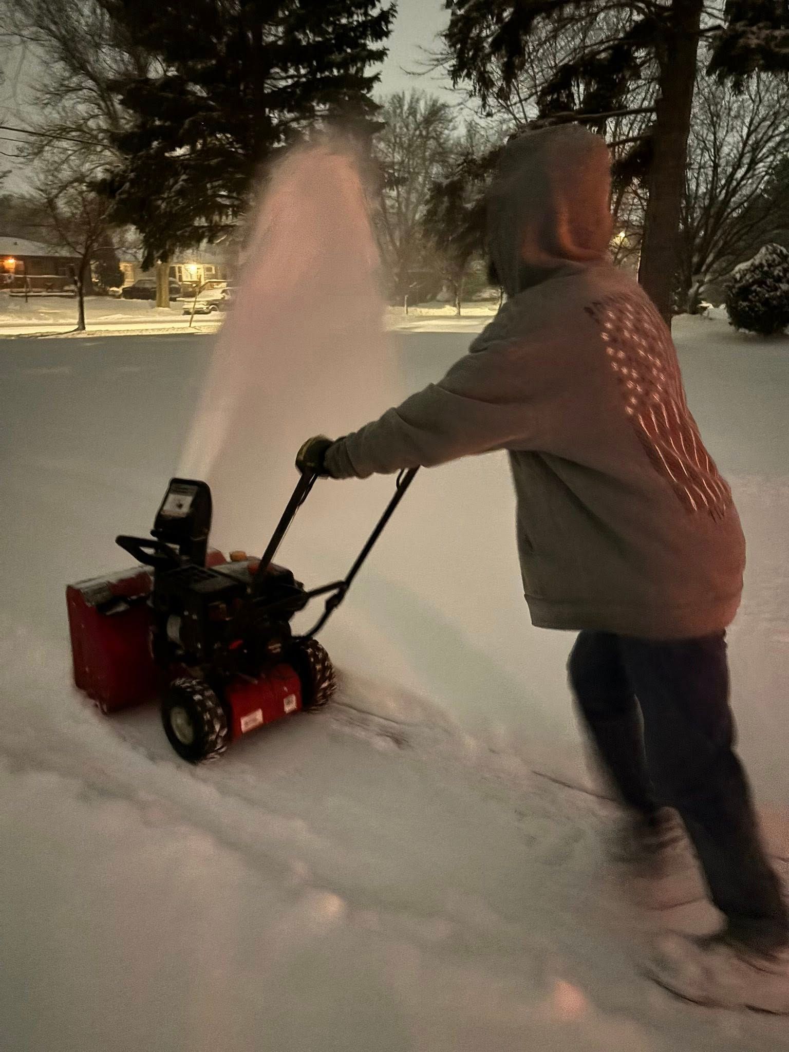 Person pushing a red snow blower through fresh snow at night, spraying snow into the air