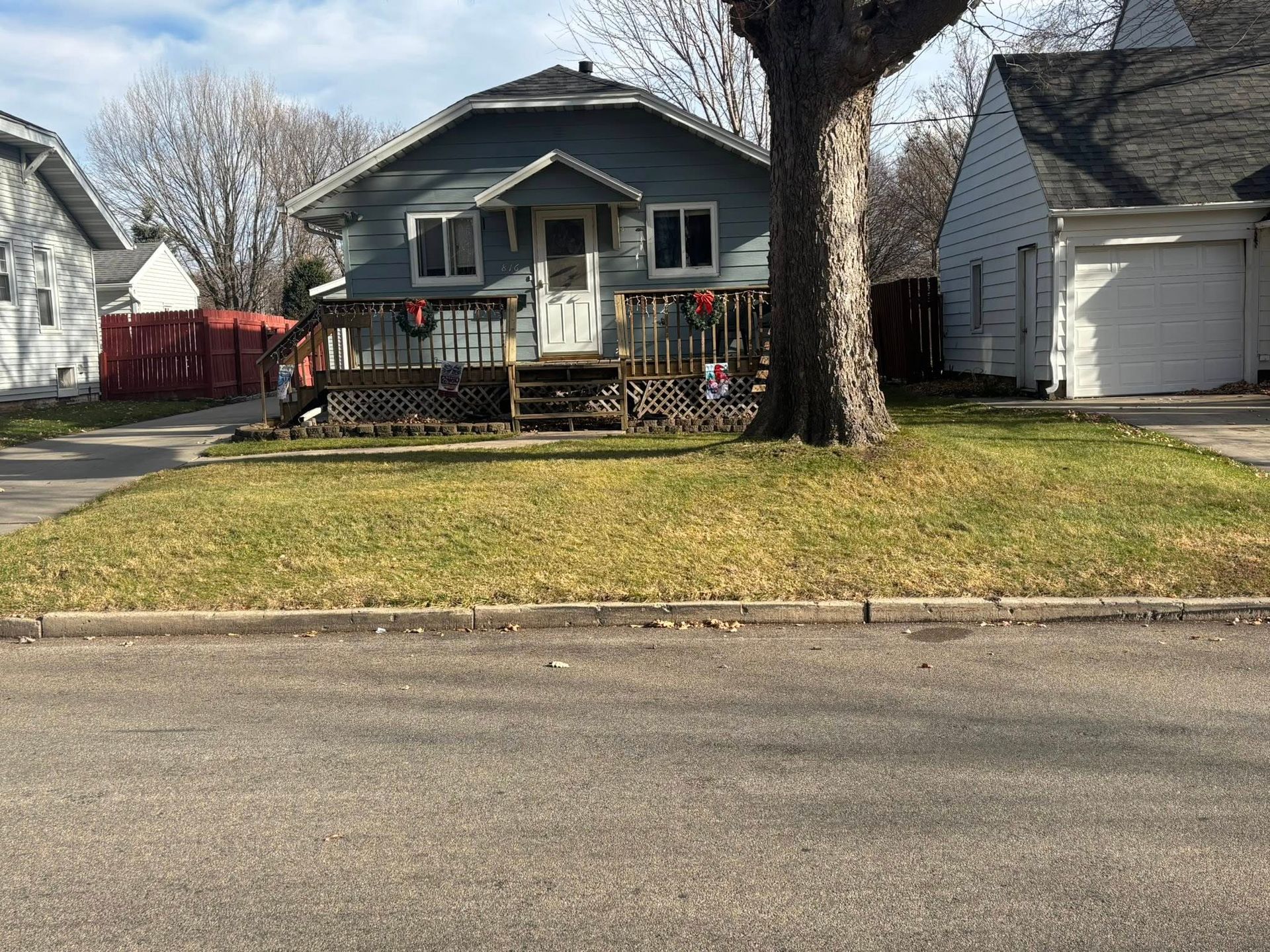 Blue house with front porch and large tree beside a street, with neighboring houses in the background