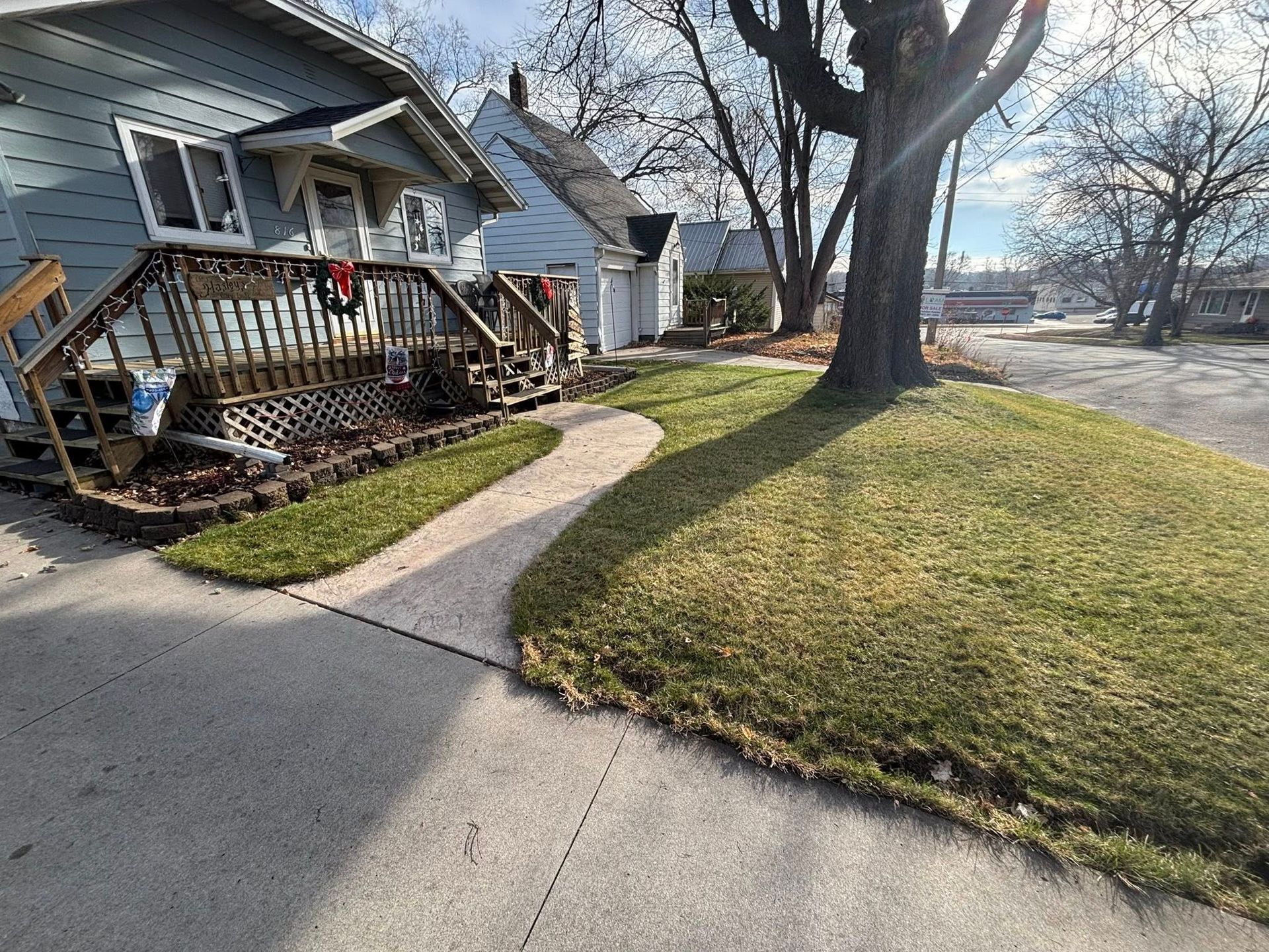 Front yard with a curved concrete walkway, small house with wooden porch, tree, and winter grass under a cloudy sky