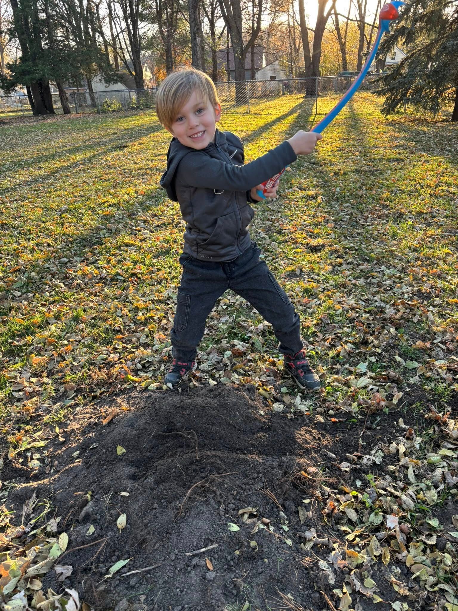 Child swinging a blue baseball bat outdoors in a leafy park, smiling beside a dirt patch.