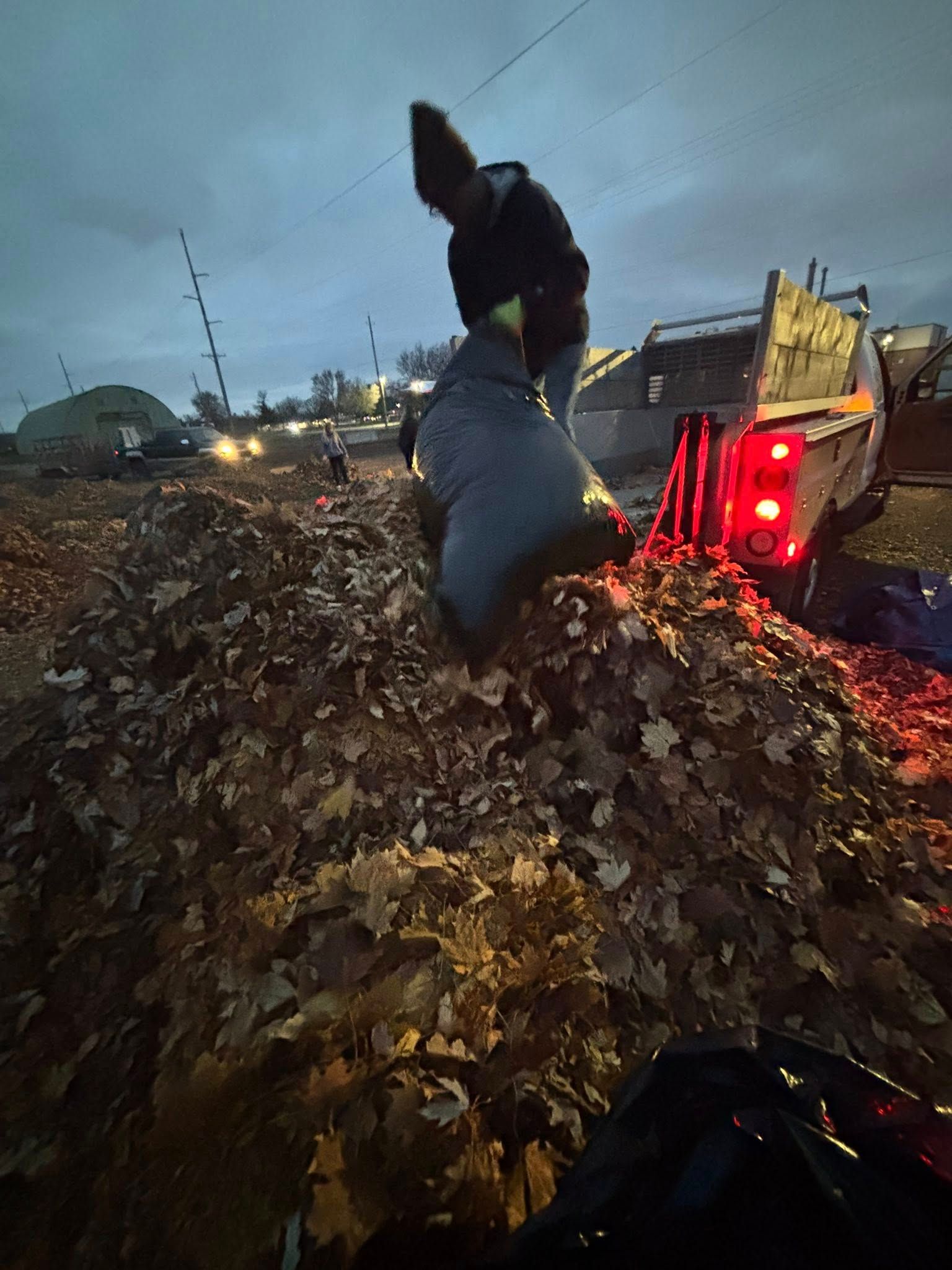 Person on a large pile of debris beside a loader at dusk, with red taillights glowing.