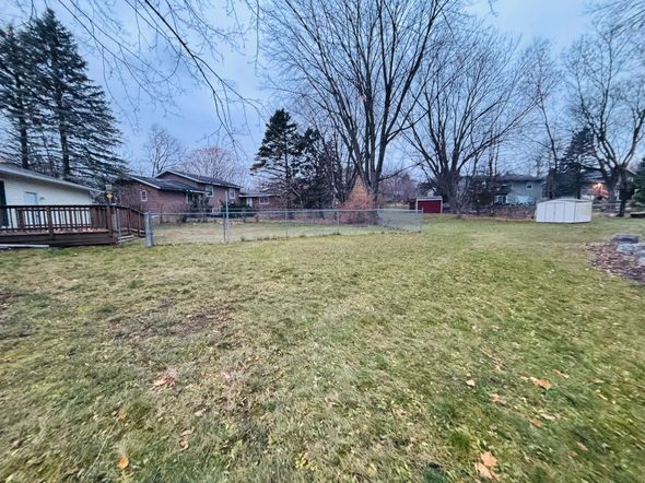 Grassy backyard with leafless trees, a wooden deck, and houses under a cloudy sky.