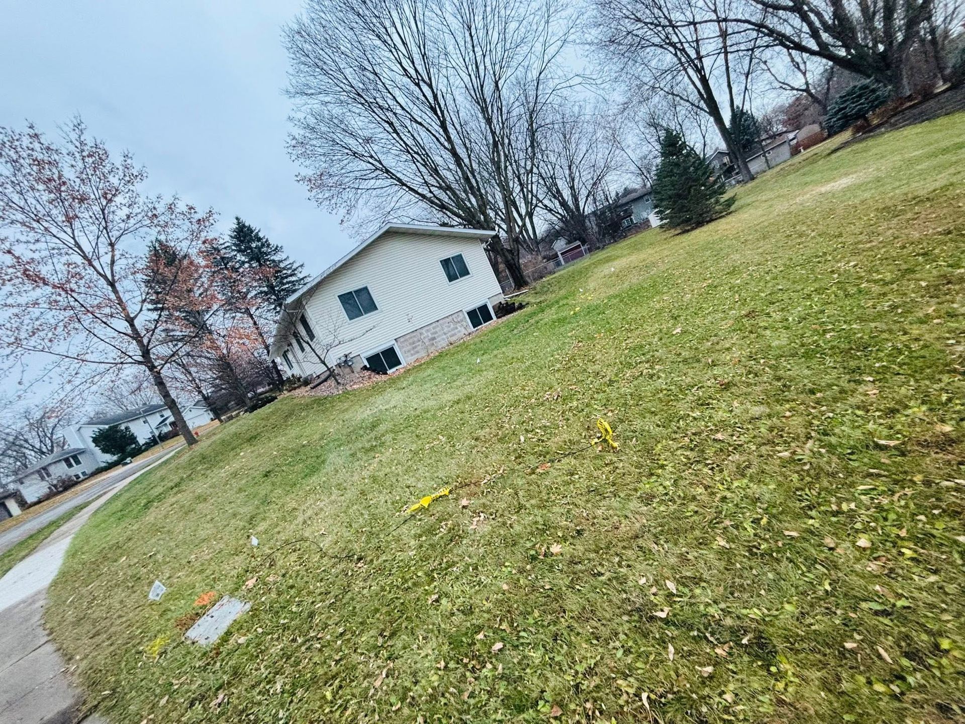Tilted view of a grassy yard with a white house, leafless trees, and a sidewalk under a cloudy sky