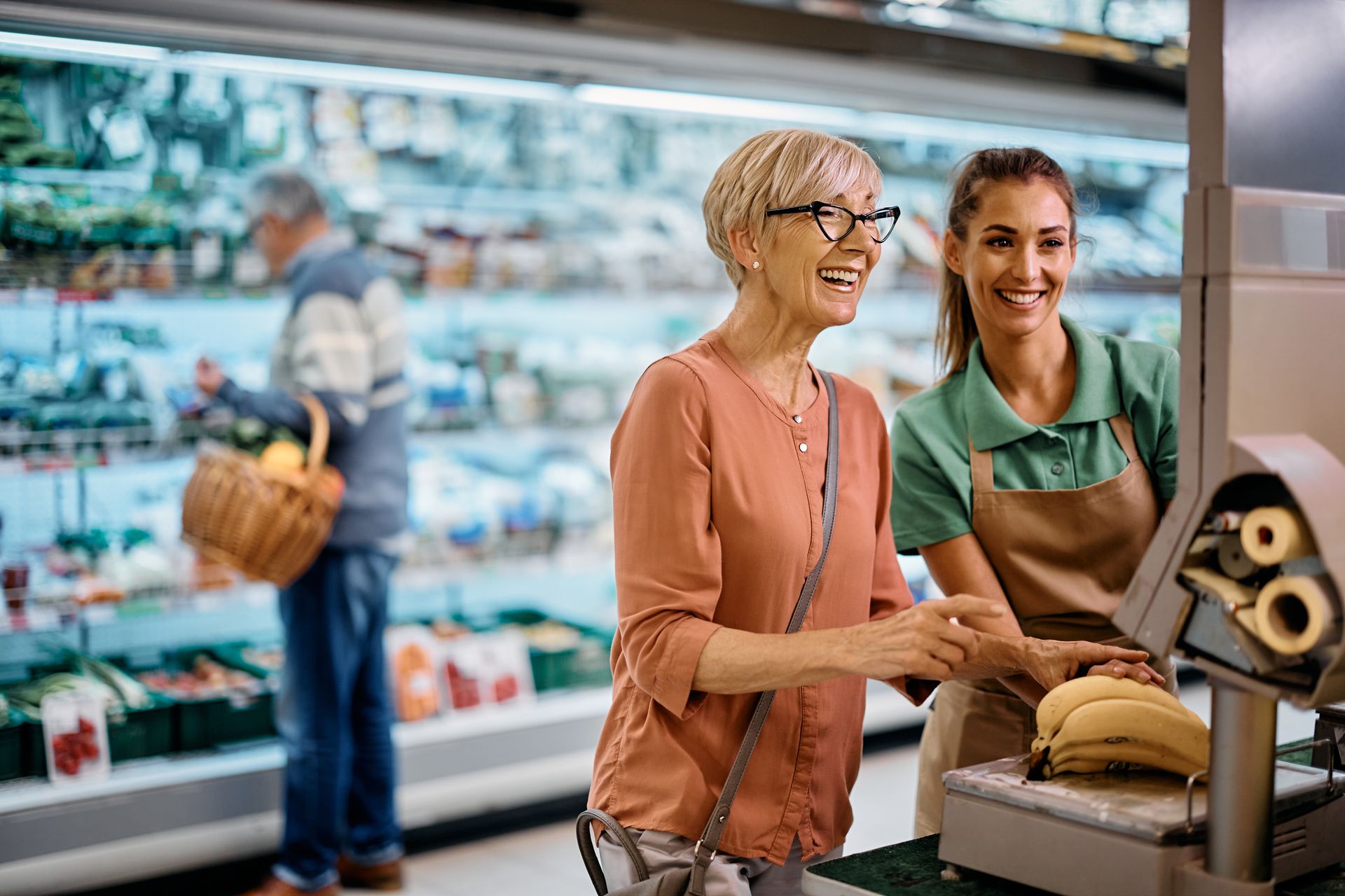 (Staff Assistance Training) A young shop assistant assists an elderly woman with weighing bananas at the supermarket scales.