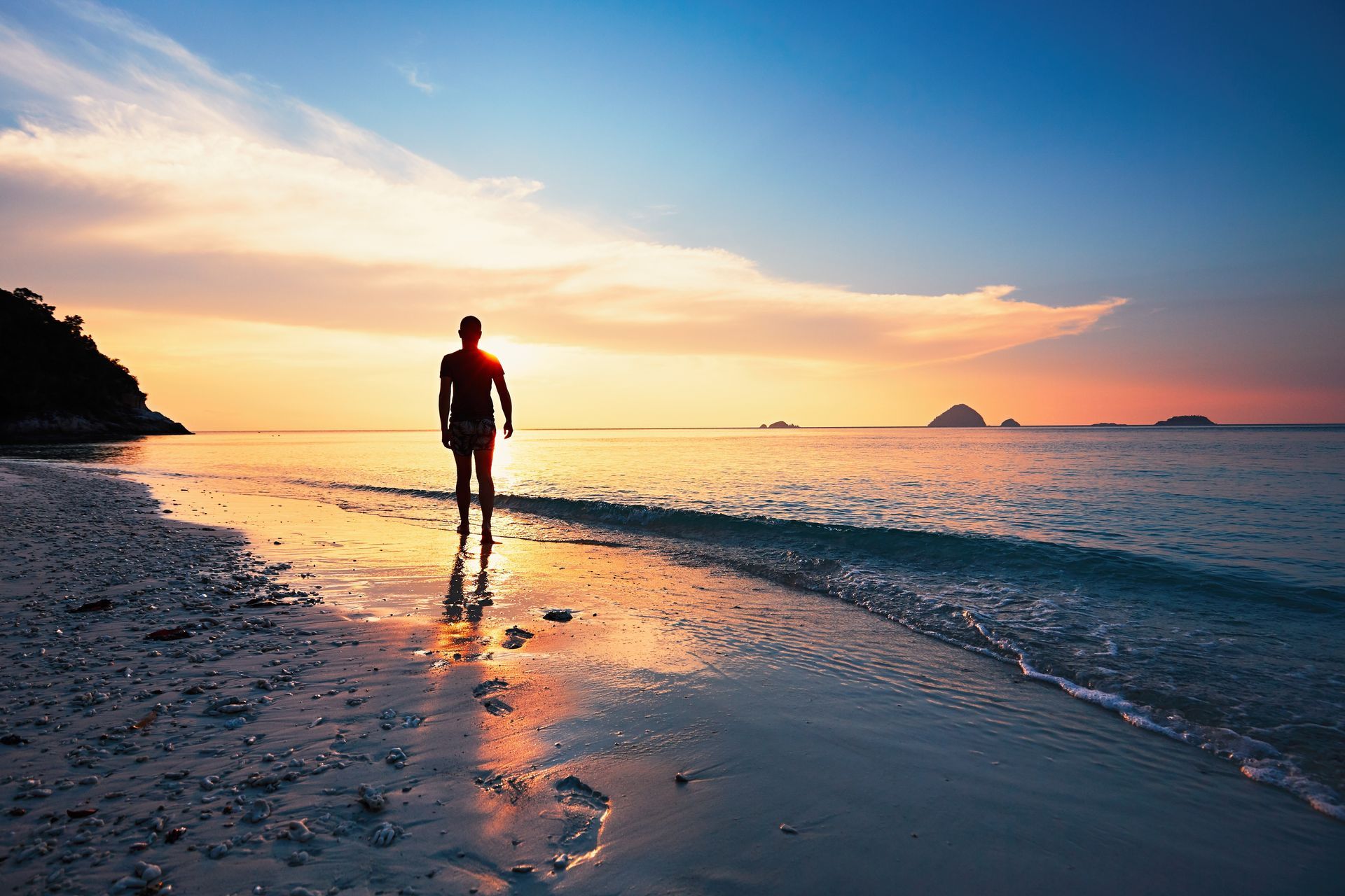 Photograph of figure walking along a beach, leaving footprints as they head towards the sunset on the horizon.