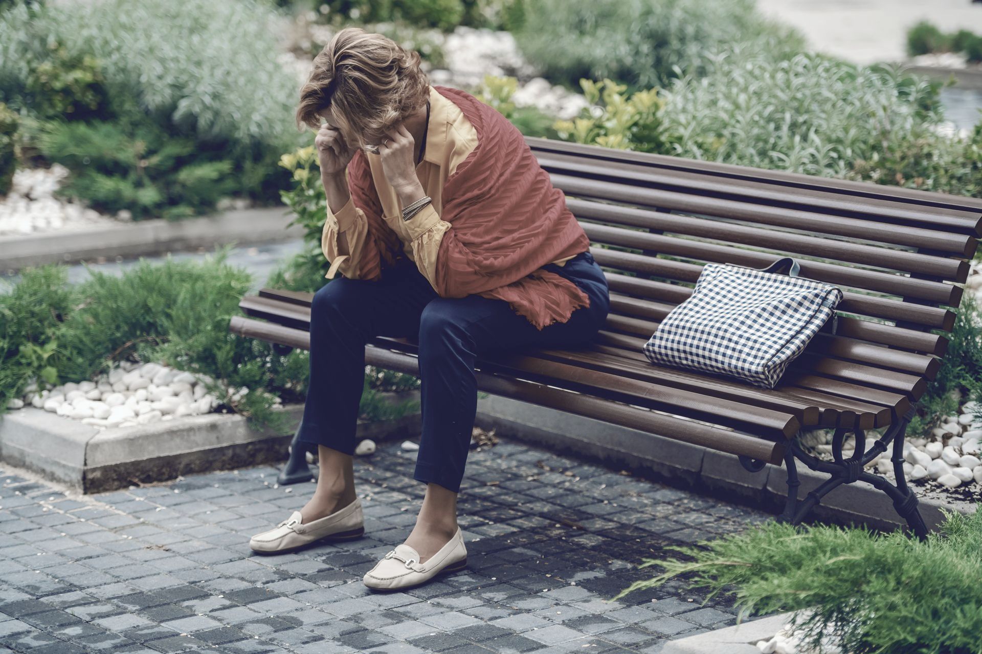Photograph of a middle-aged lady sat alone on a park bench holding her head in her hands.