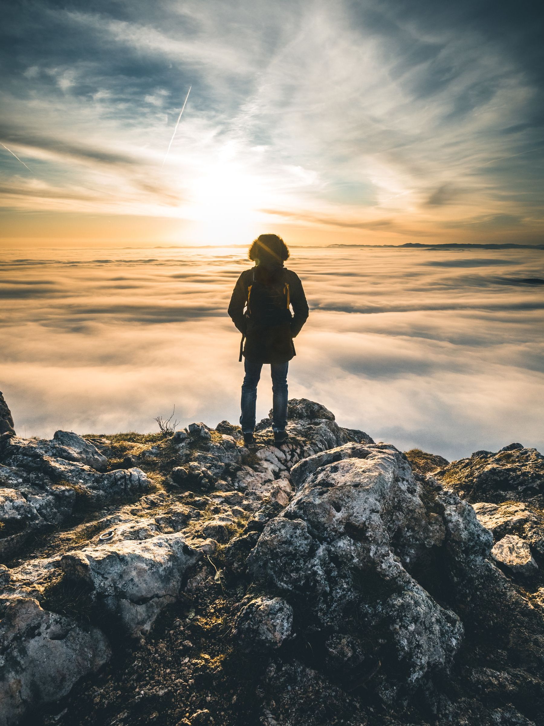 A hiker stands at the peak of a snowy mountain, gazing into a blazing sunset and a blanket of clouds below. 