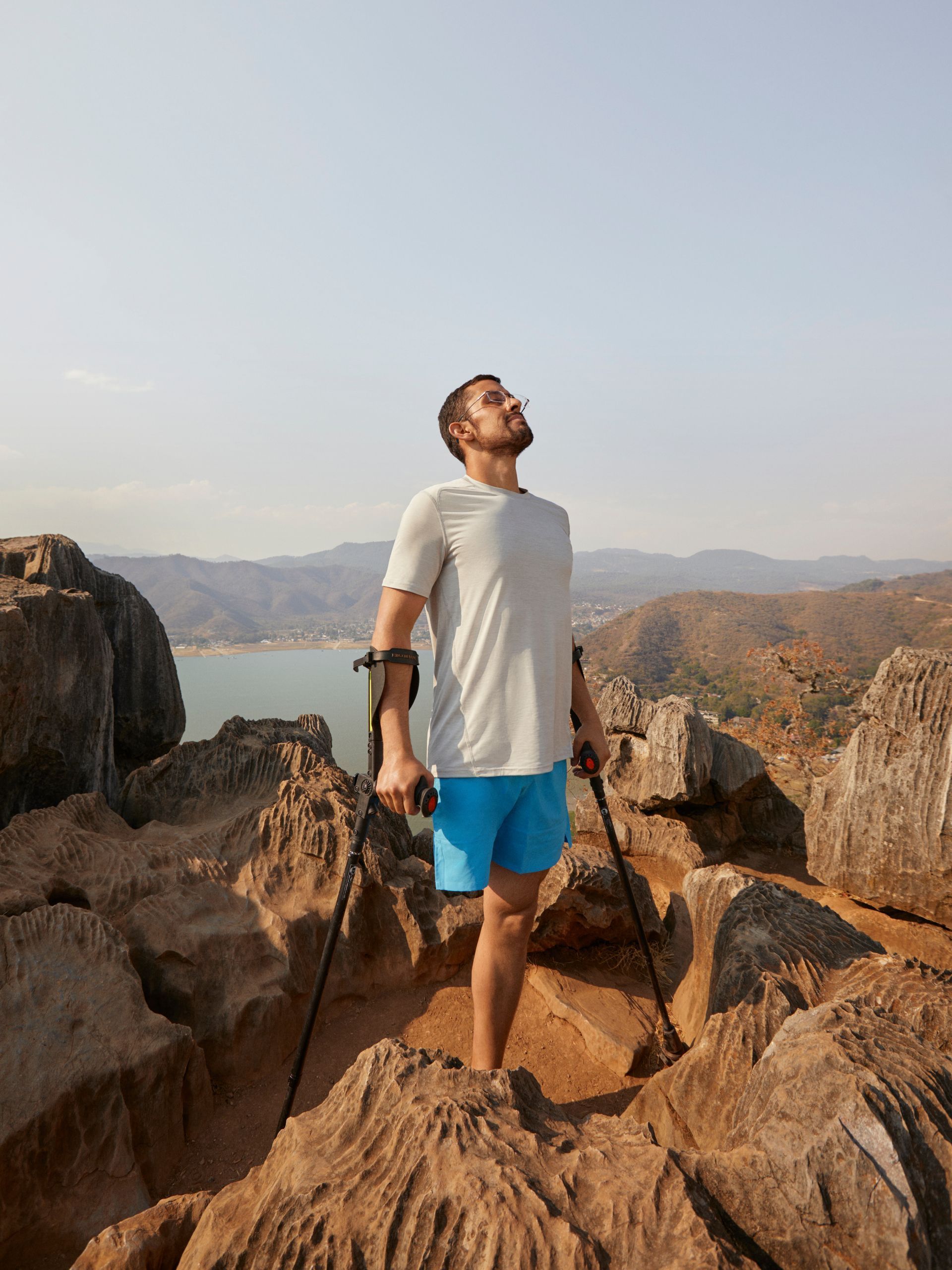 On an high, rocky outcrop, a man with crutches and a lower limb amputation breathes in the fresh air after a long climb.