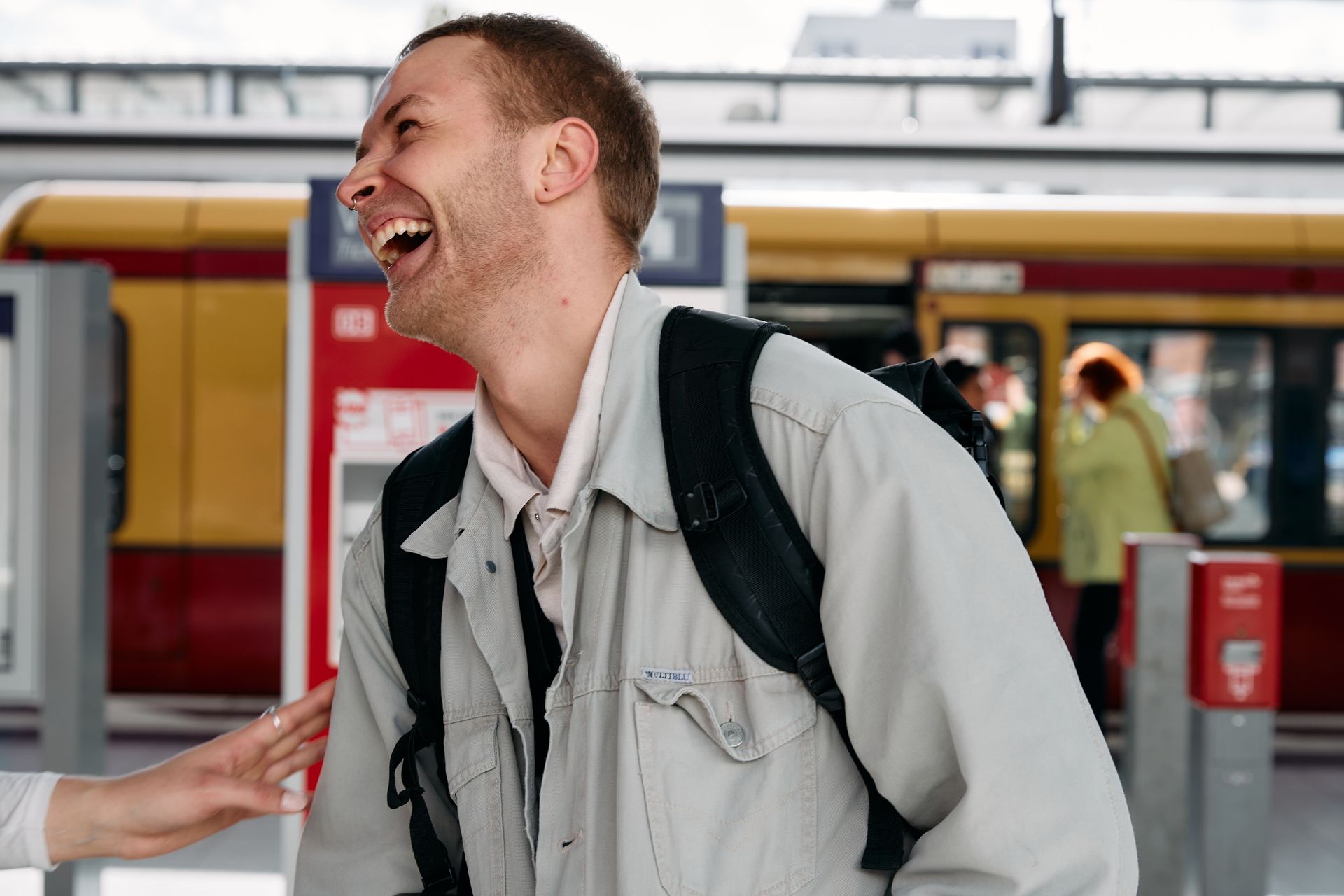 A young man laughs as he buys train tickets. In the background, a train awaits its passengers in a busy station.