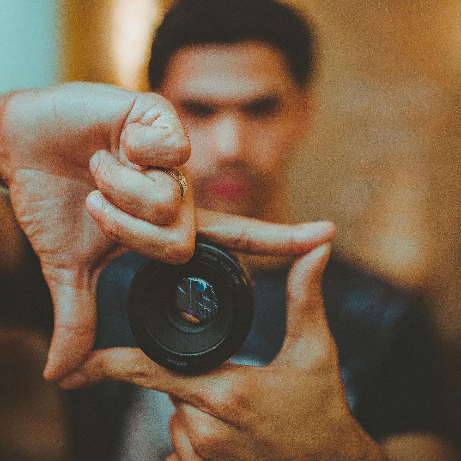 A man holds a camera lens in front of his face, framing his view with his hands. The background is blurred, drawing focus to the lens, which captures a sharp, selective portion of the scene. This represents how our perception is filtered—seeing only what aligns with our expectations while the bigger picture remains out of focus.