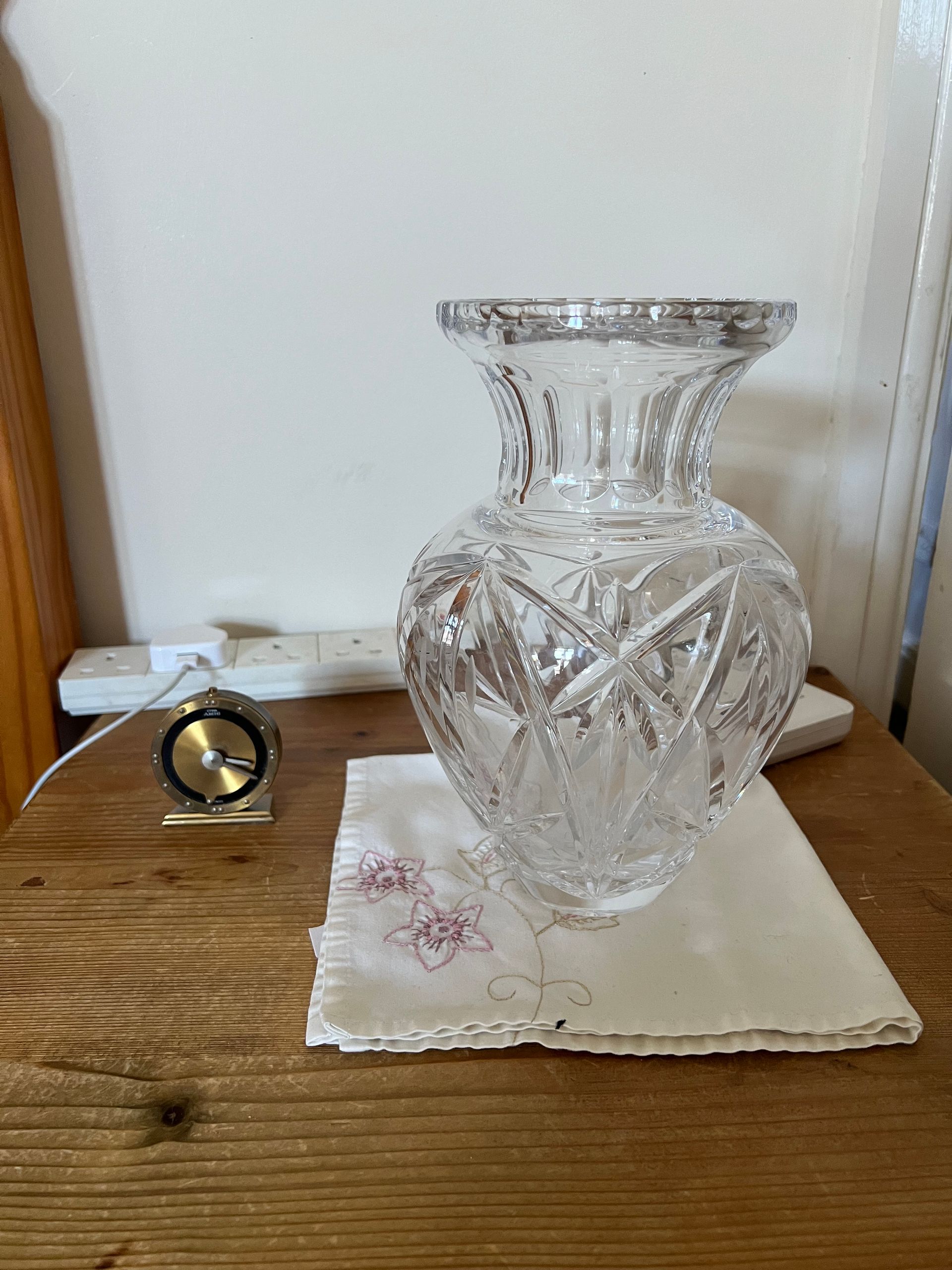 A mechanical alarm clock and glase vase on a bedside table.