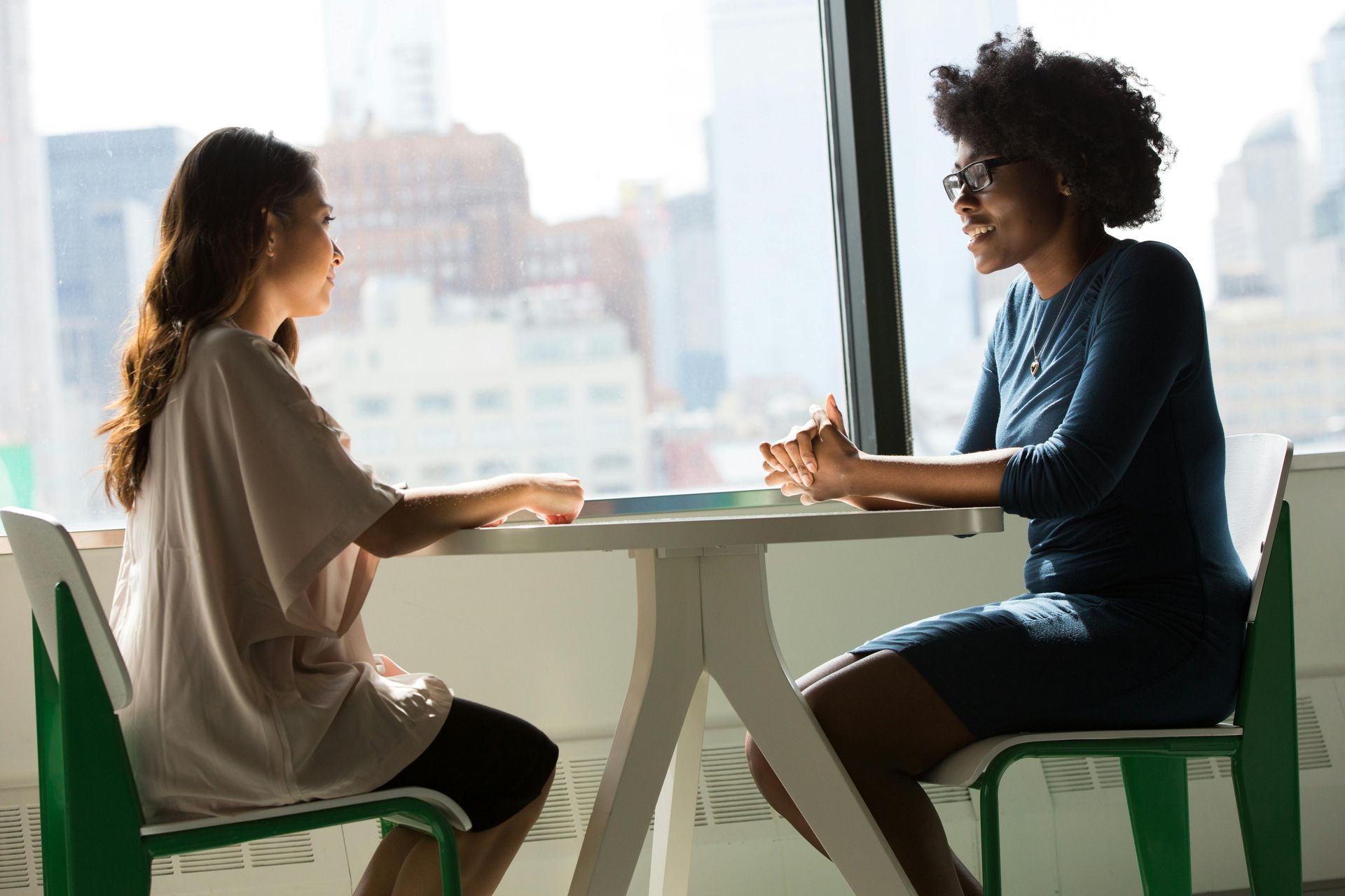 Two women sit across from each other at a white table by a large window in a bright, modern setting. Sunlight streams in, casting soft shadows. The woman on the left, wearing a light blouse and dark shorts, faces the woman on the right, who wears a fitted navy dress and glasses. Both appear engaged and relaxed, hands rested, sharing a thoughtful moment of conversation against a blurred urban skyline.