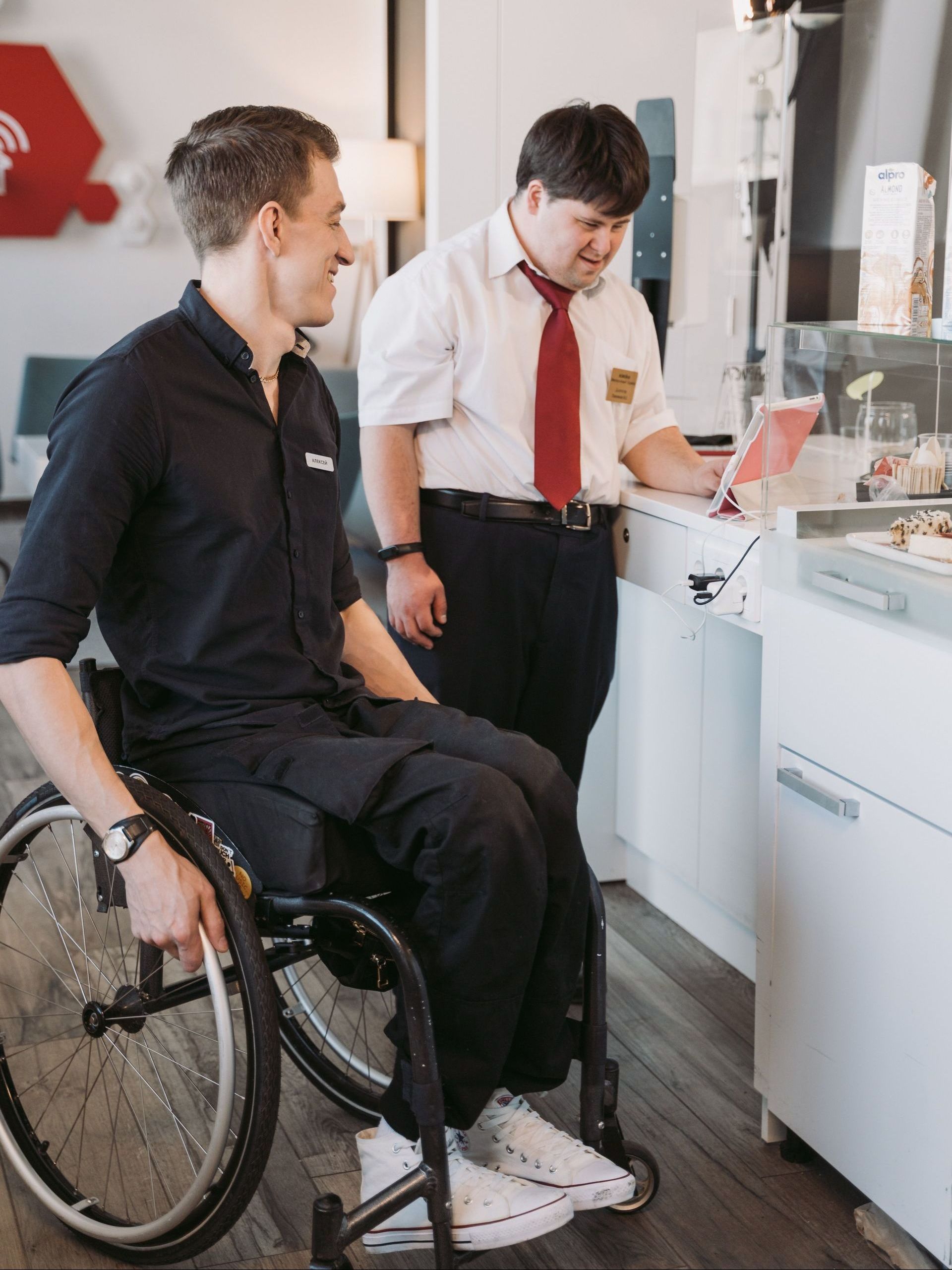 A male supervisor with Down Syndrome and a cafe worker in a wheelchair looking on a computer behind the counter.