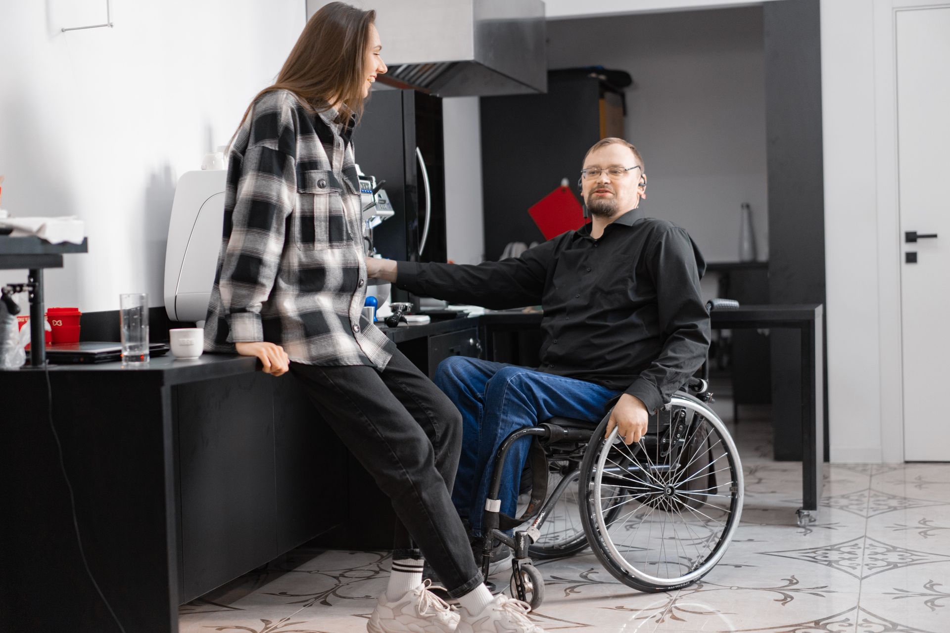 An older man with hearing loss and a mobility impairment chats with a casually dressed young woman at a work coffee station.
