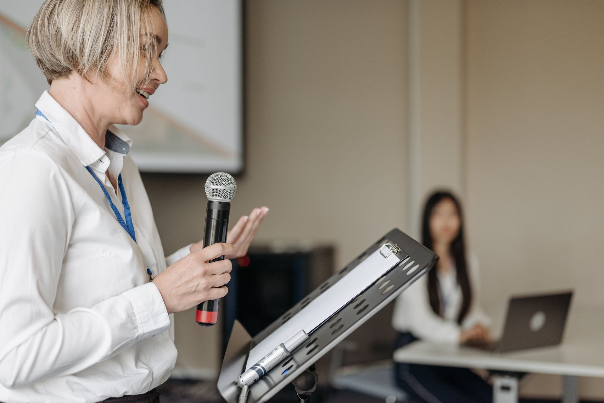 A mature woman in smart attire who stands behind a metal podium overcomes her nerves and speaks to a conference via a microphone.