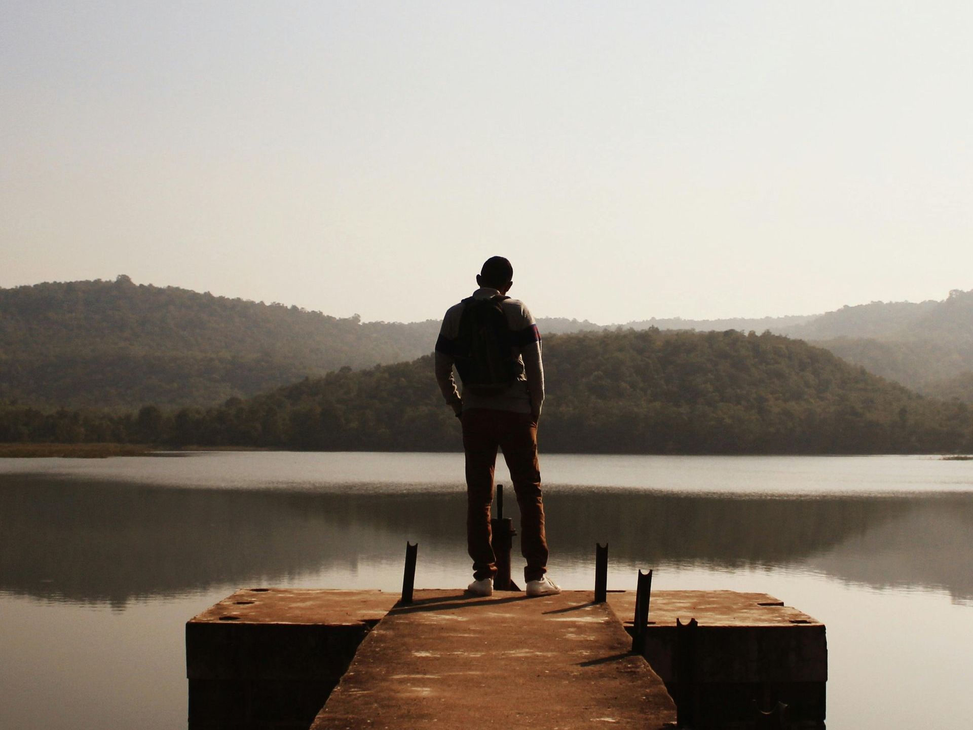 A solitary silhouette stands on a pier, gazing at a calm lake during sunset, evoking serenity and reflection. 
