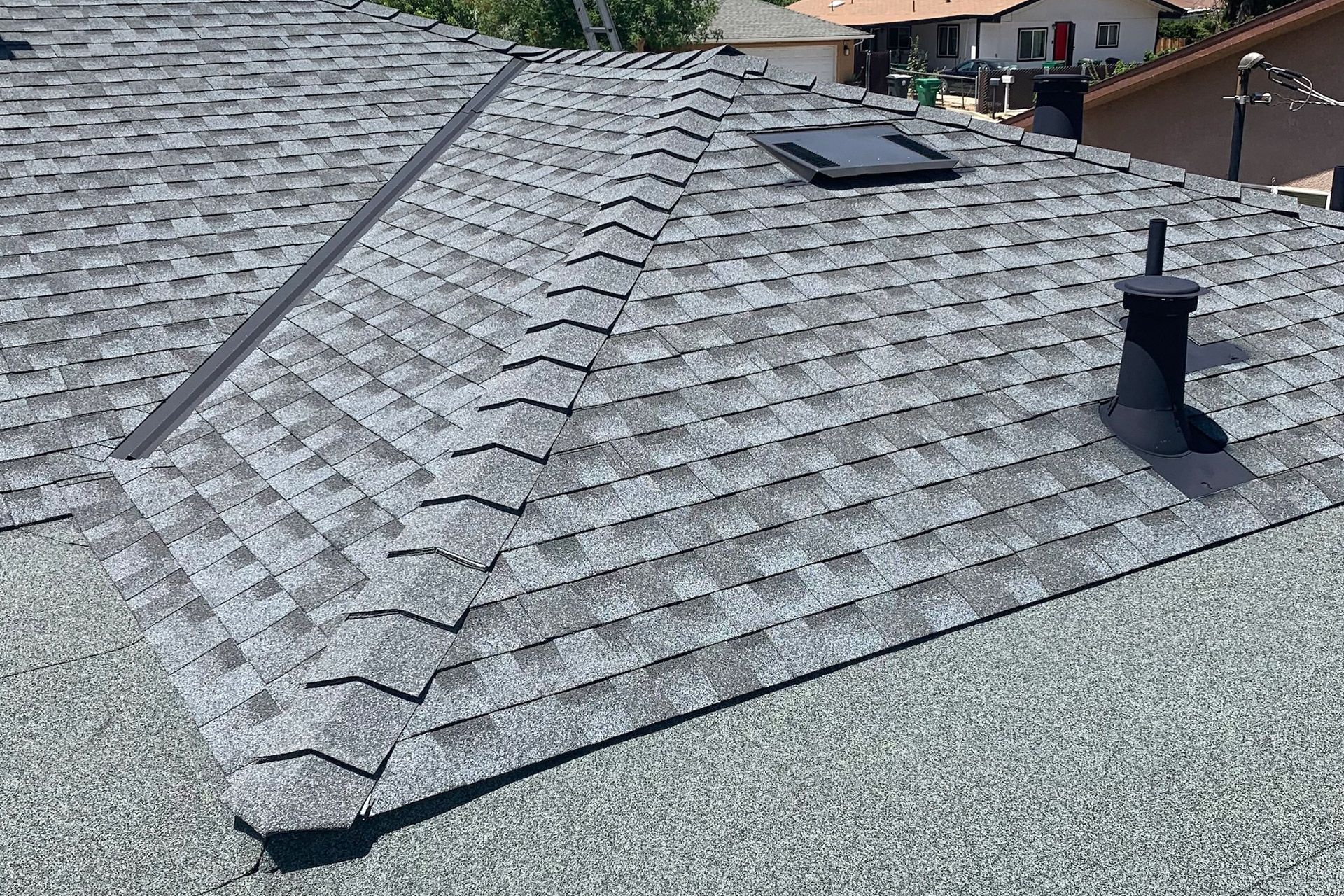 Gray asphalt shingle roof with a vent, chimney, and metal flashing on a sunny day.