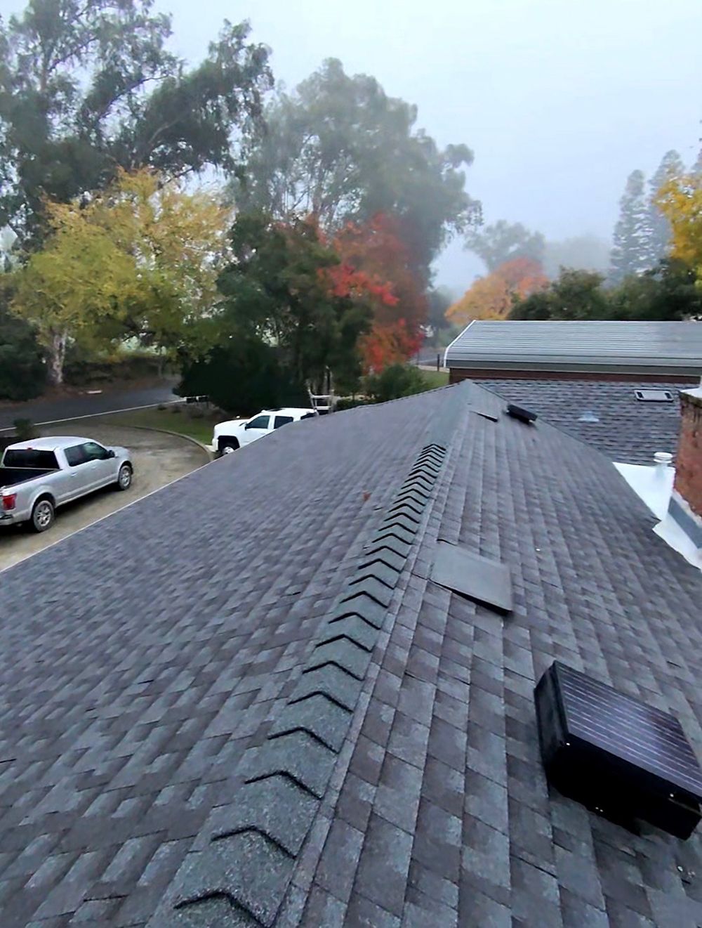 Gray shingle roof with a central ridge vent, overlooking a foggy scene with fall foliage and parked trucks.