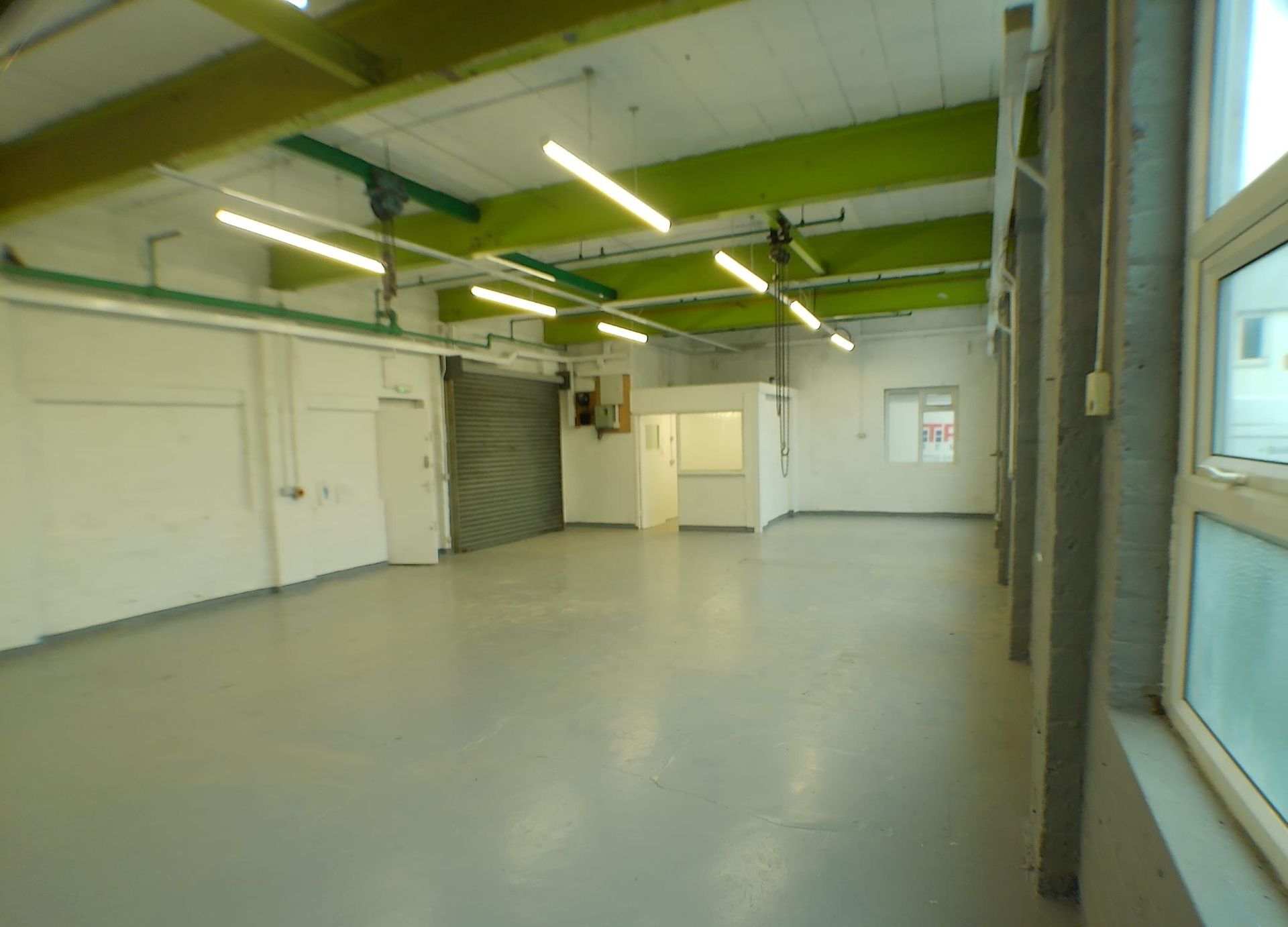 Empty industrial room with gray floor, white walls, green ceiling beams, and fluorescent lights