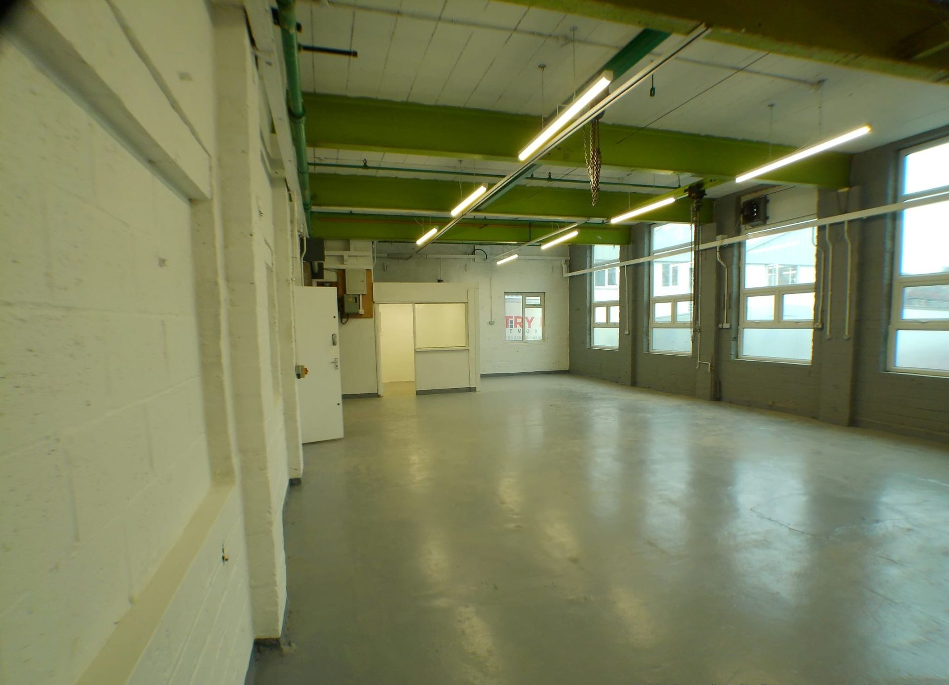 Empty industrial room with gray floor, green ceiling beams, and large windows on the right.