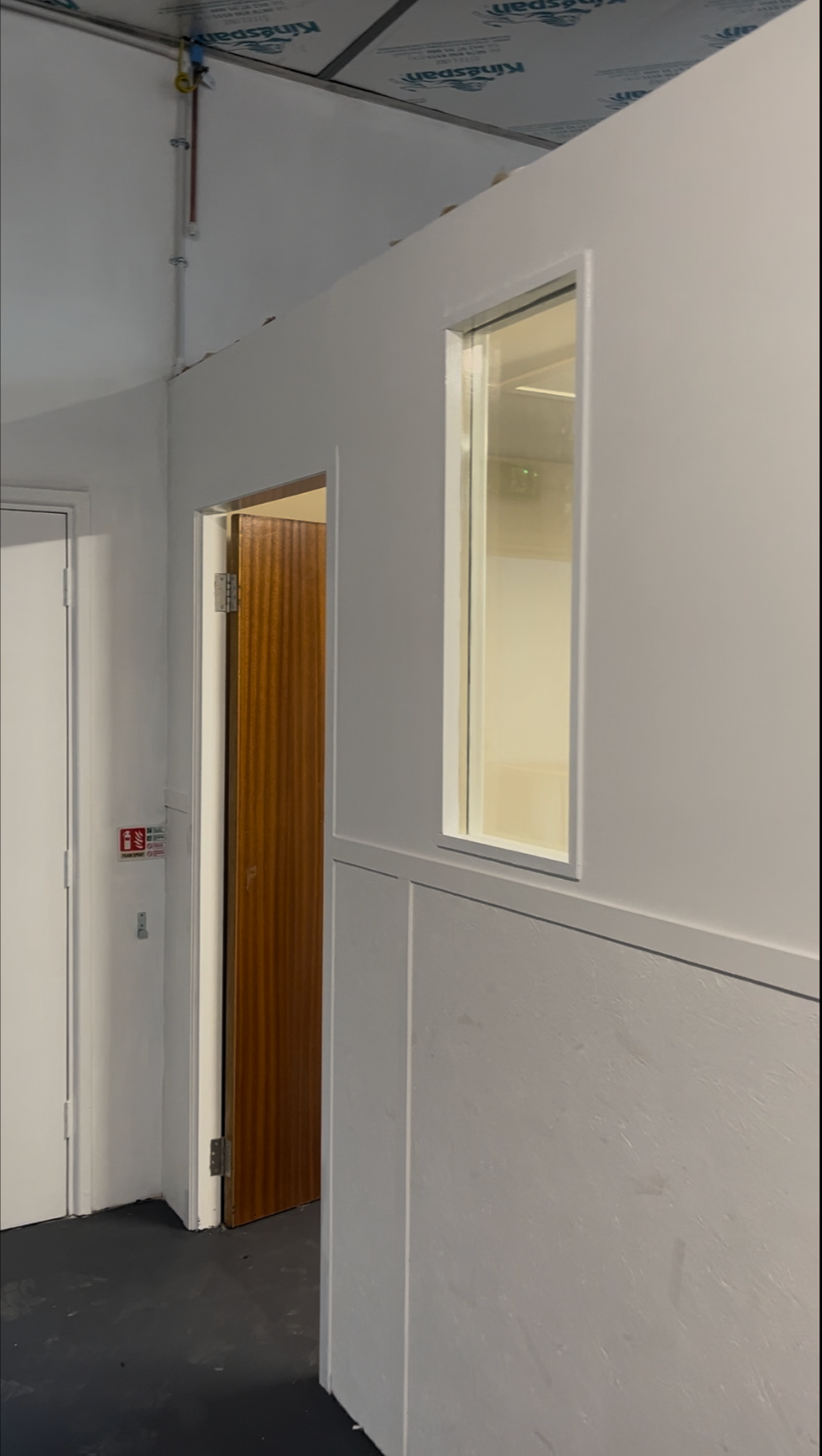 White hallway with a wooden door, narrow window, and drop ceiling tiles.