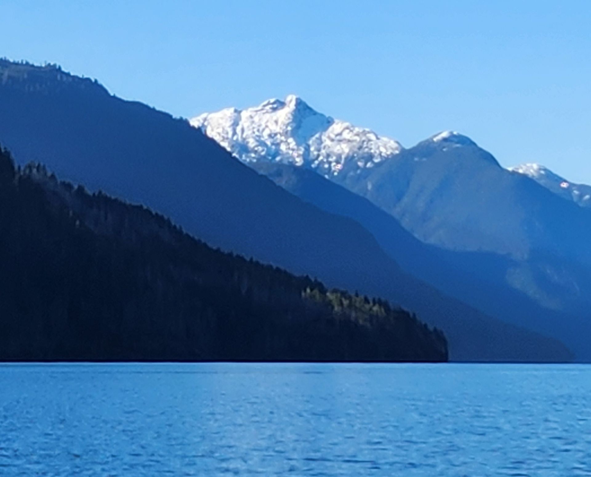 Snowy mountain peaks above a blue lake, with dark forested hills in the foreground.