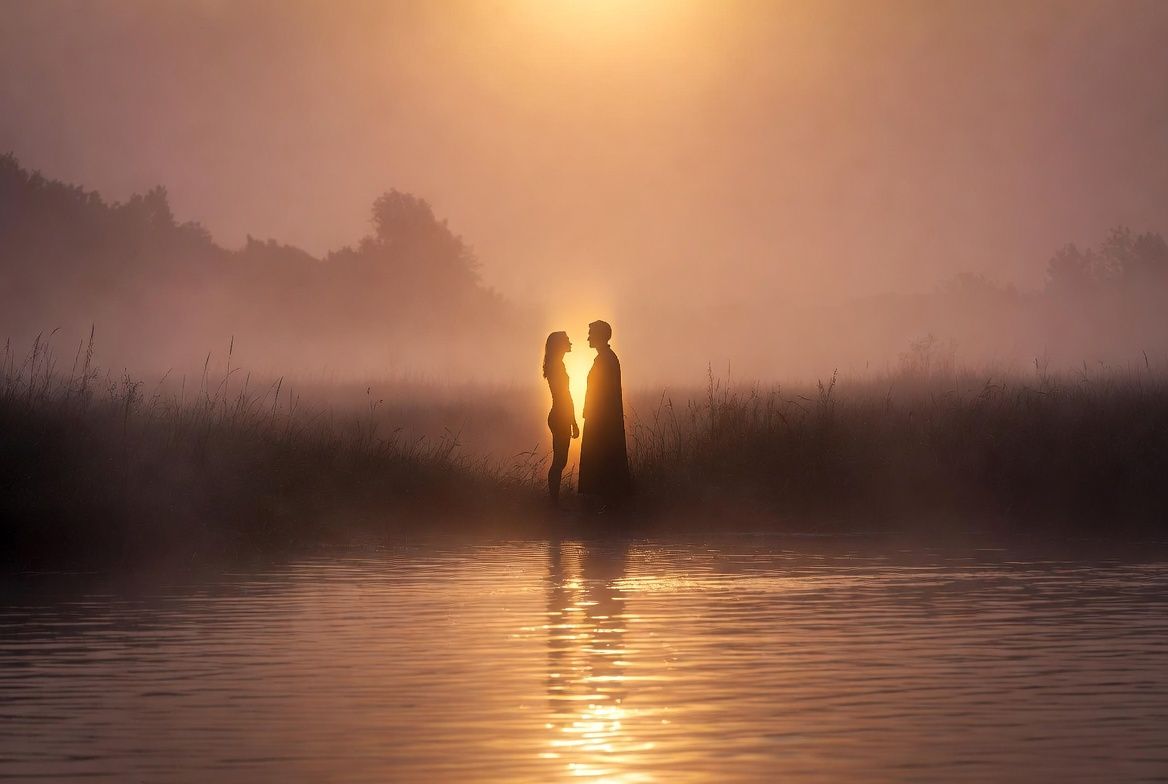 silhouetted woman and man facing one another standing in tall grass at the edge of a pond at sunset