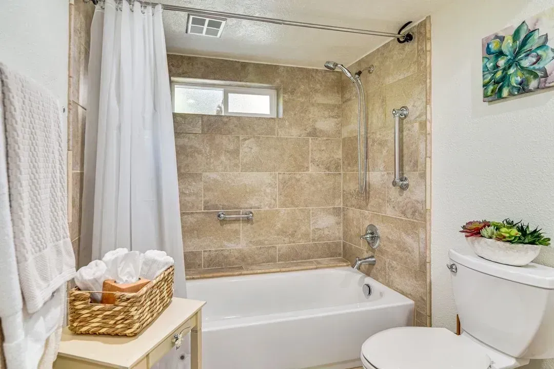 Beige tiled bathroom with a bathtub, shower curtain, towel bar, and toilet beside a small table.